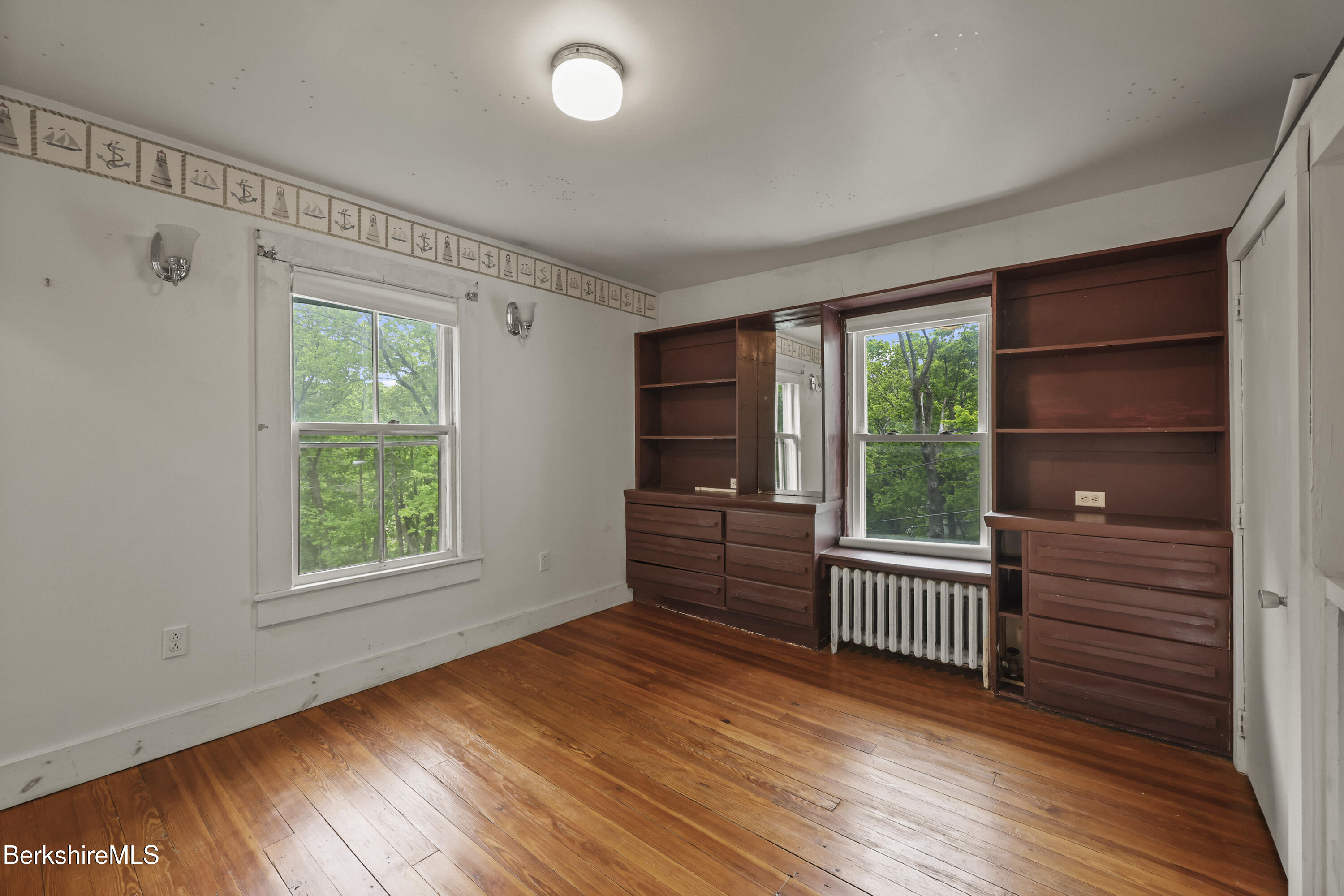 Empty room with polished hardwood floors, built-in wooden shelving and drawers, and two windows overlooking trees.