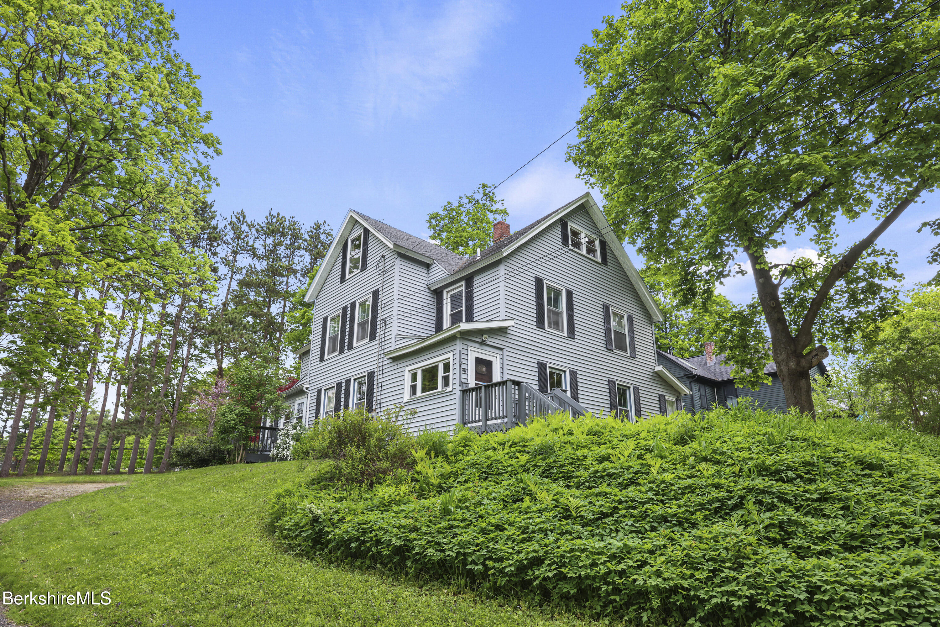 Tall gray house with dark shutters and a small porch above a lush green lawn and shrubs.