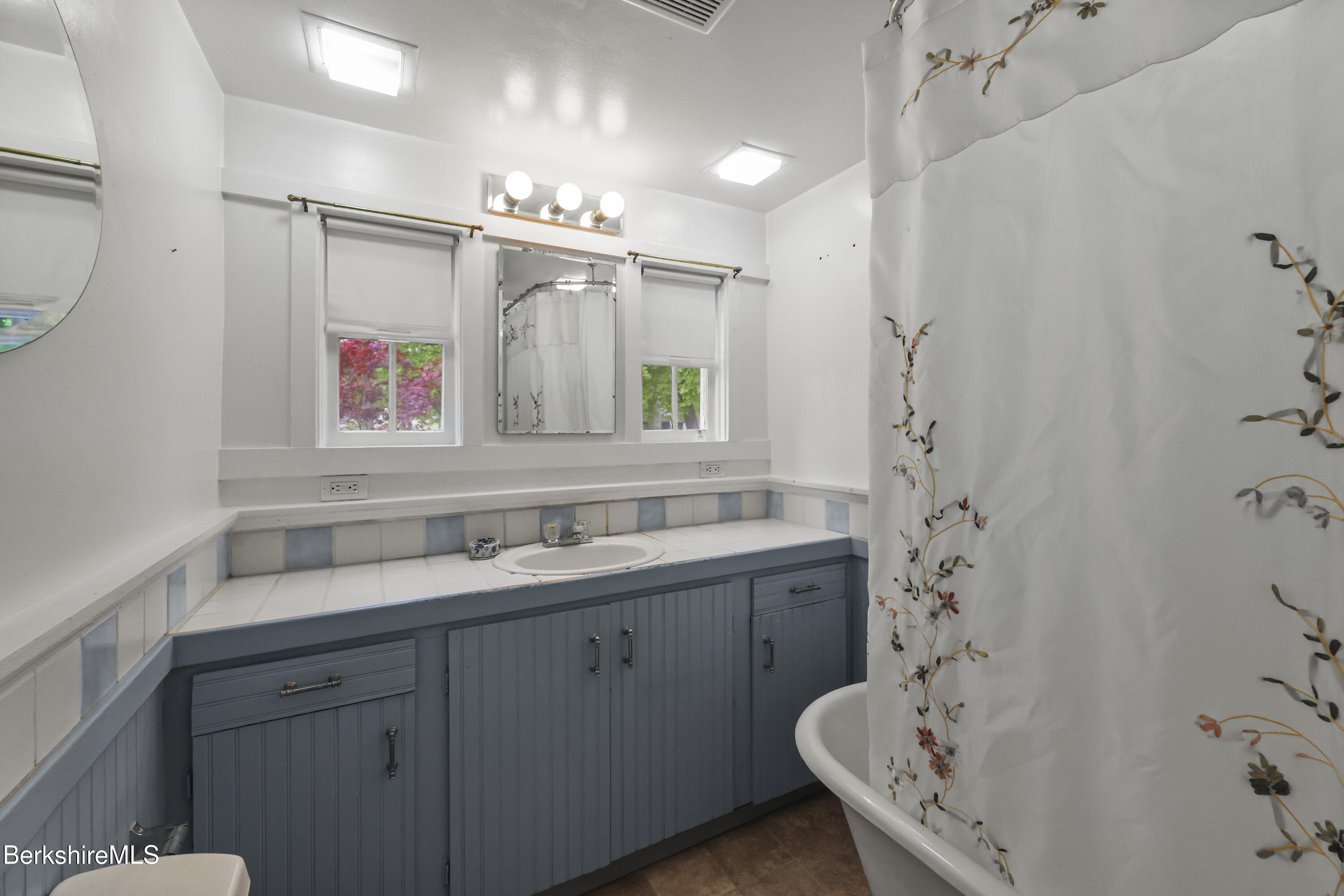 Bathroom with a blue paneled vanity, tiled counter, two windows, and floral shower curtain.