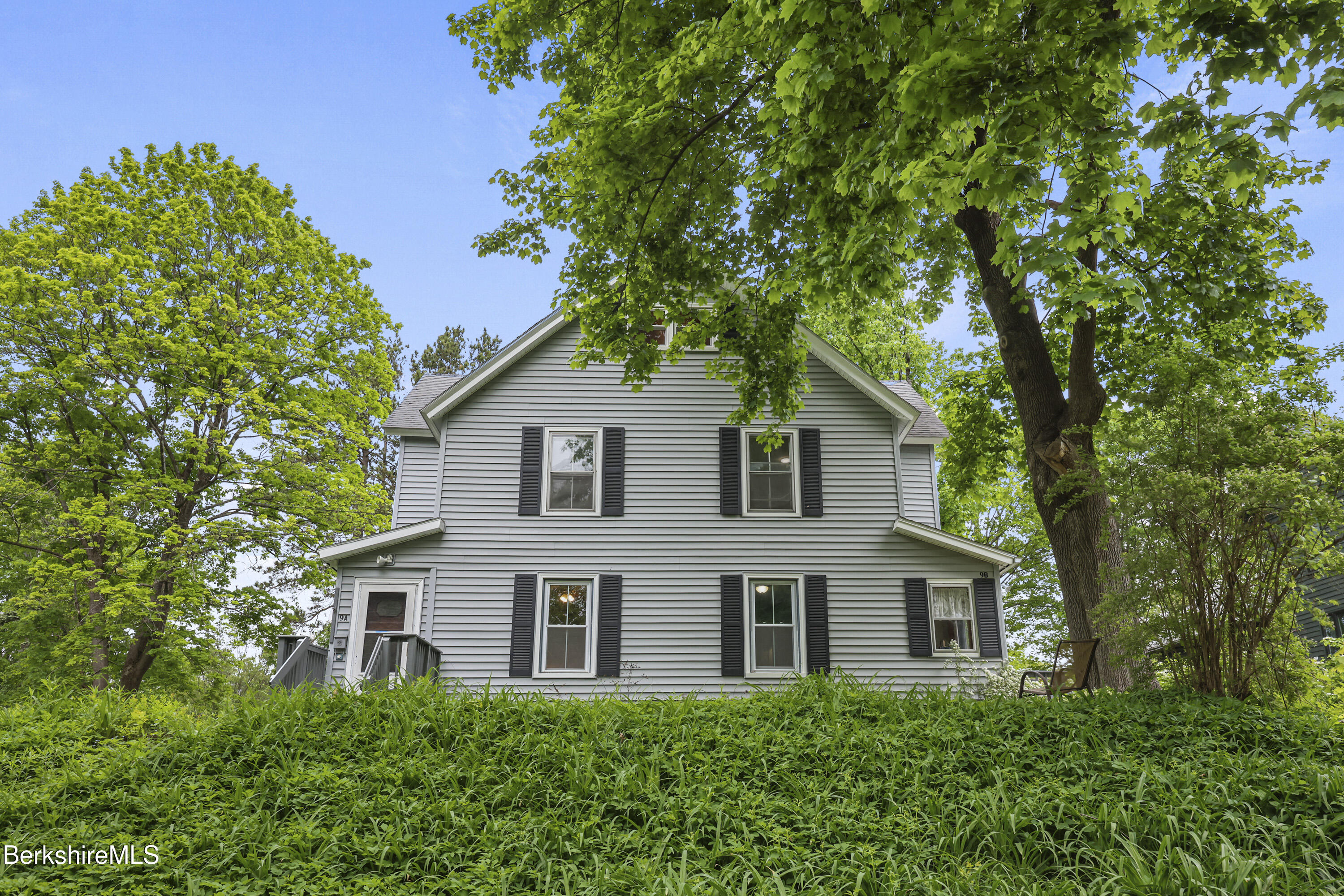Gray two-story house with black shutters, framed by mature leafy trees and an overgrown lawn.