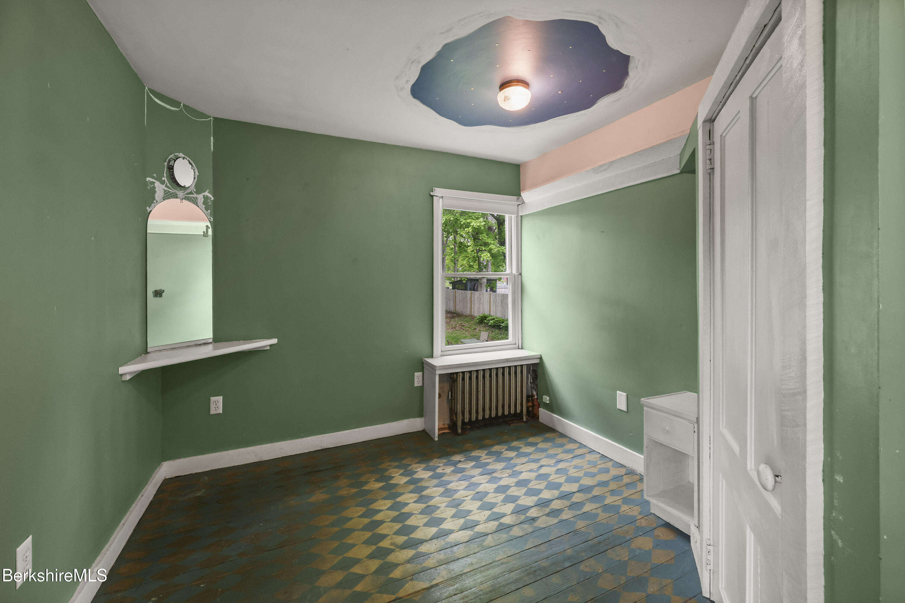 Empty green room with a blue-and-tan checkerboard floor, wall mirror, and window above a radiator.