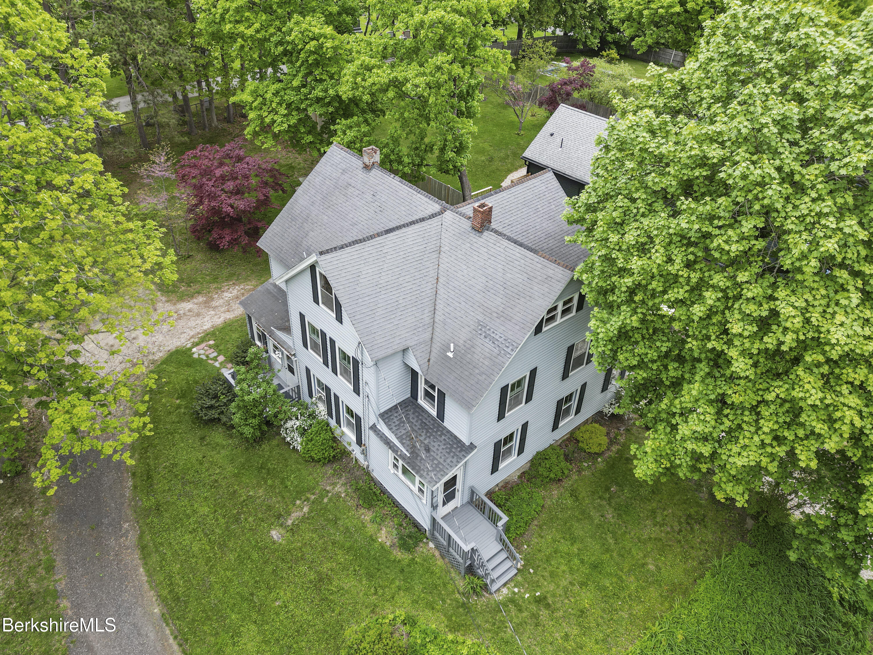 Aerial view of a gray-roofed house surrounded by mature trees, with a rear deck and gravel driveway.