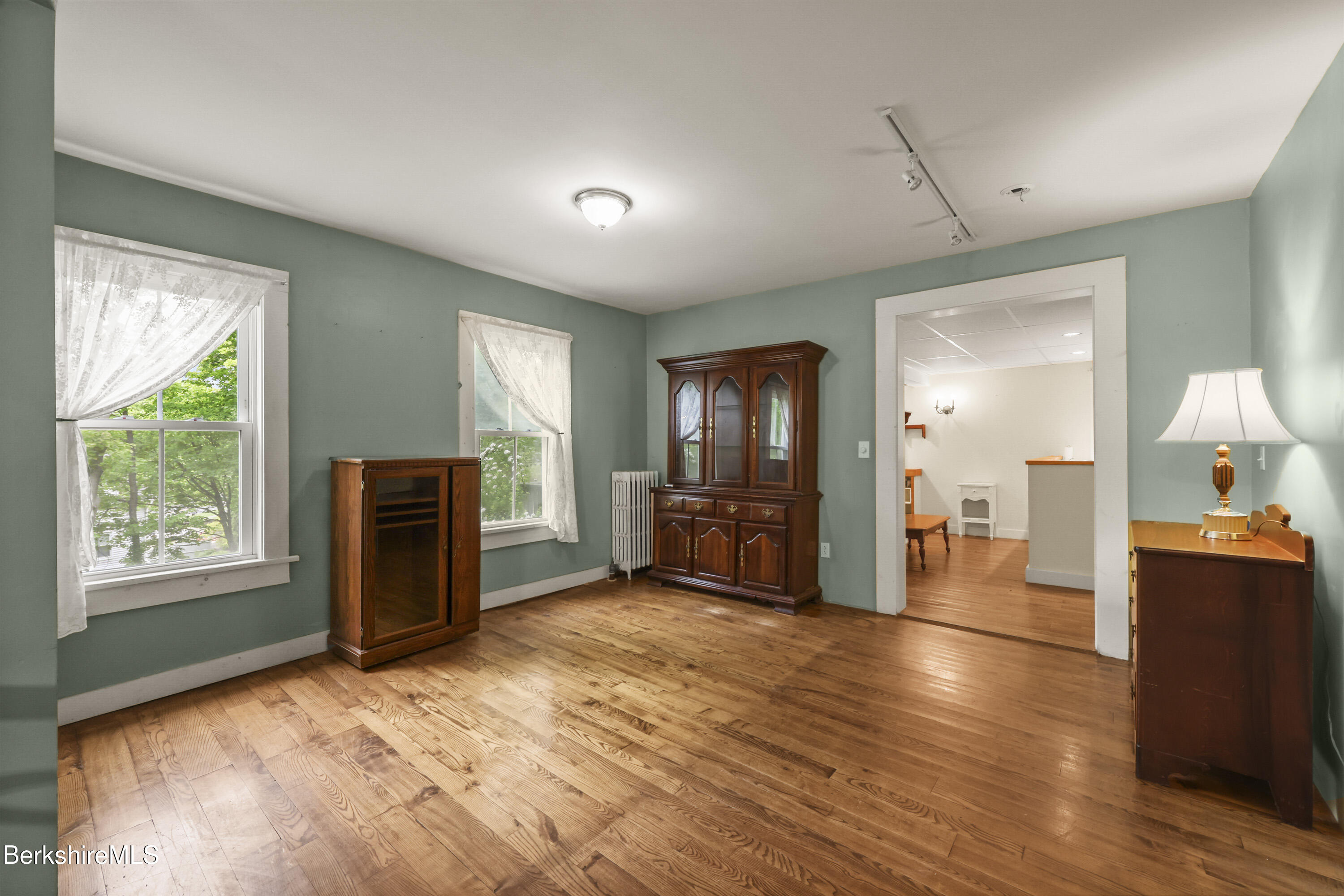 Furnished room with wide-plank wood floors, sage-green walls, and lace-curtained windows.