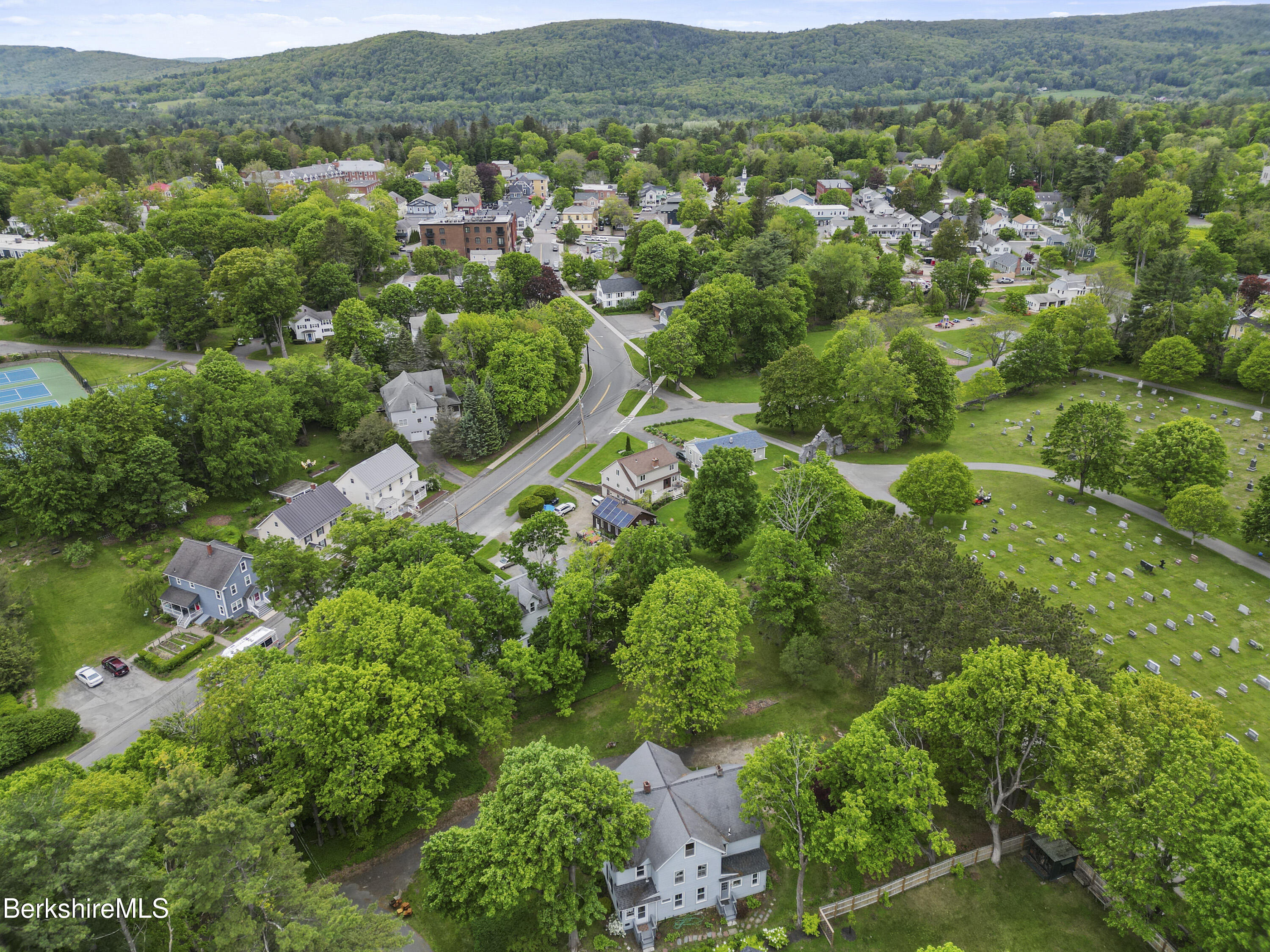 Gray multi-story house among mature trees, with a cemetery and forested hills nearby.
