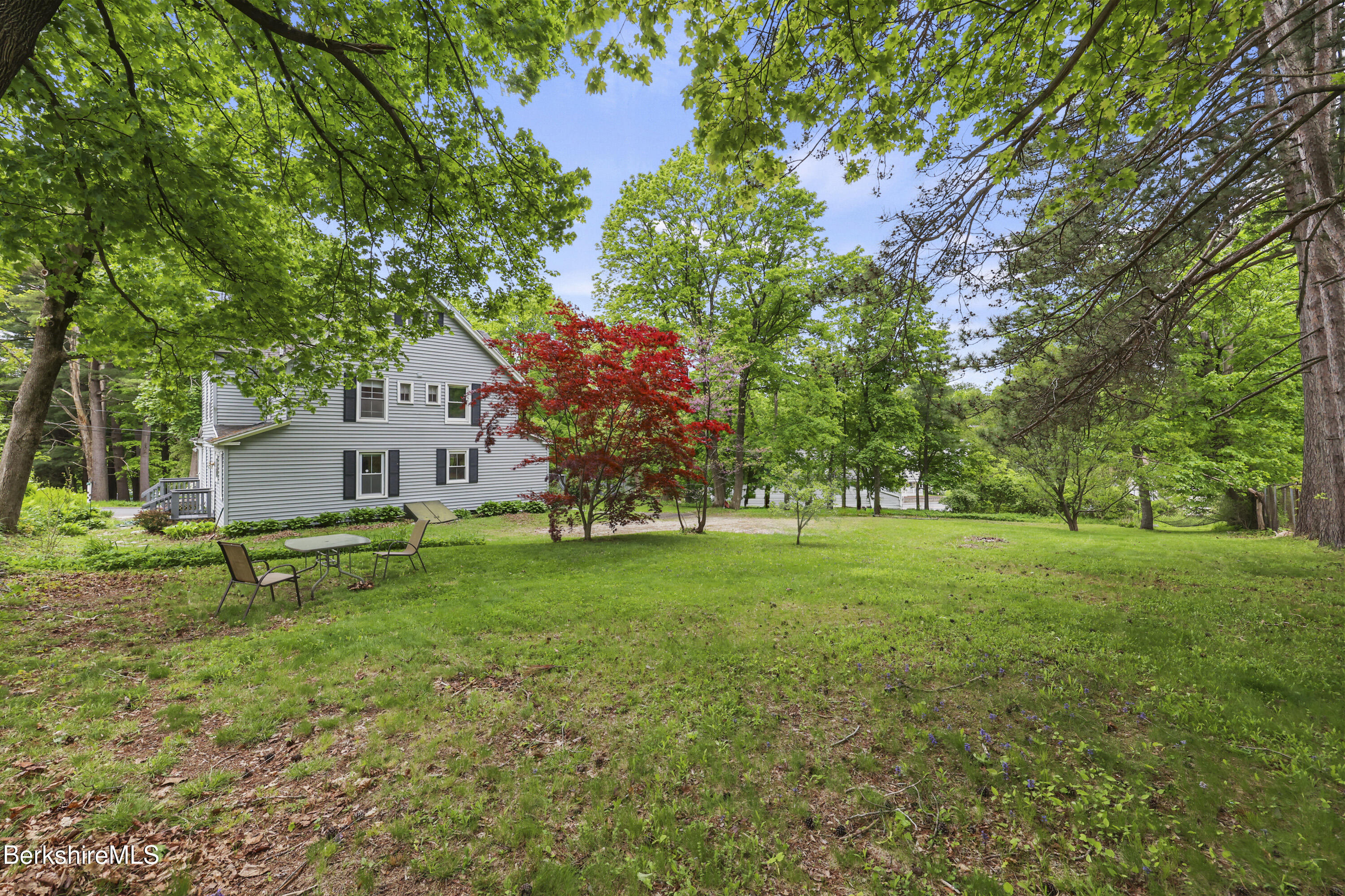 Gray-sided house among mature trees, with a broad lawn and red-leaf ornamental tree.