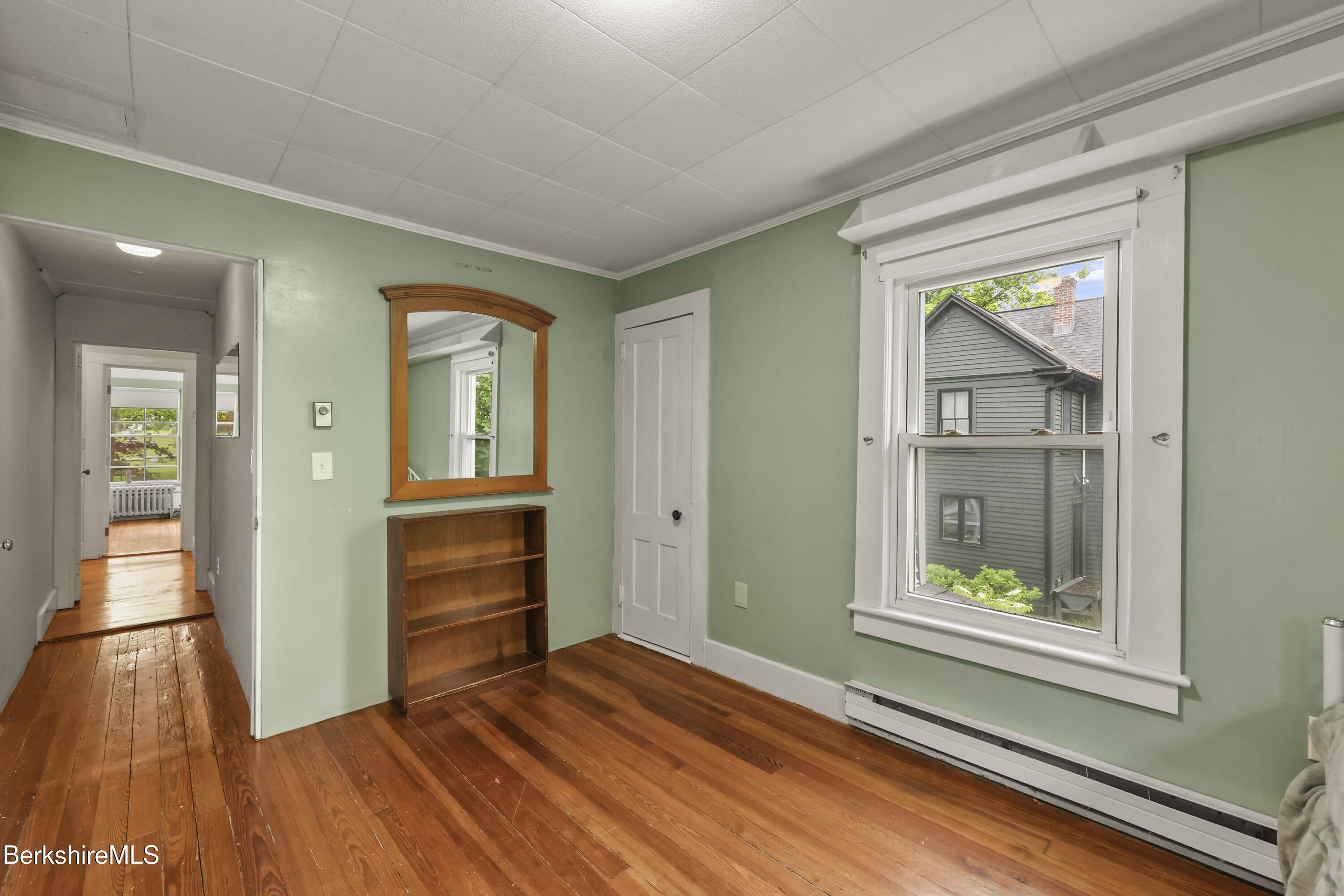 Green-walled room with wide-plank hardwood floors, a large window, and a wooden bookcase beneath a mirror.