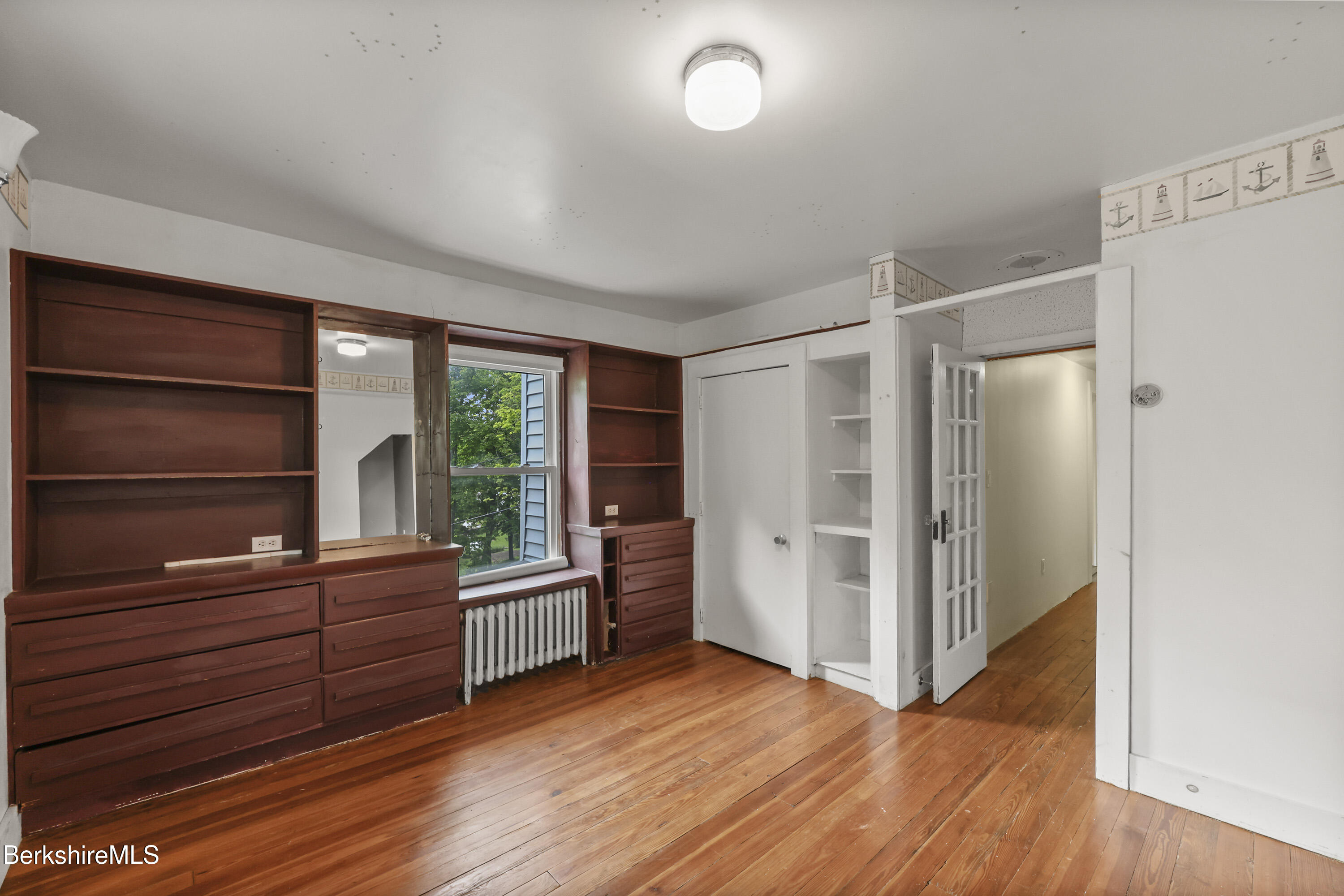 Unfurnished bedroom with built-in reddish-brown shelves, hardwood floors, and a window overlooking trees.