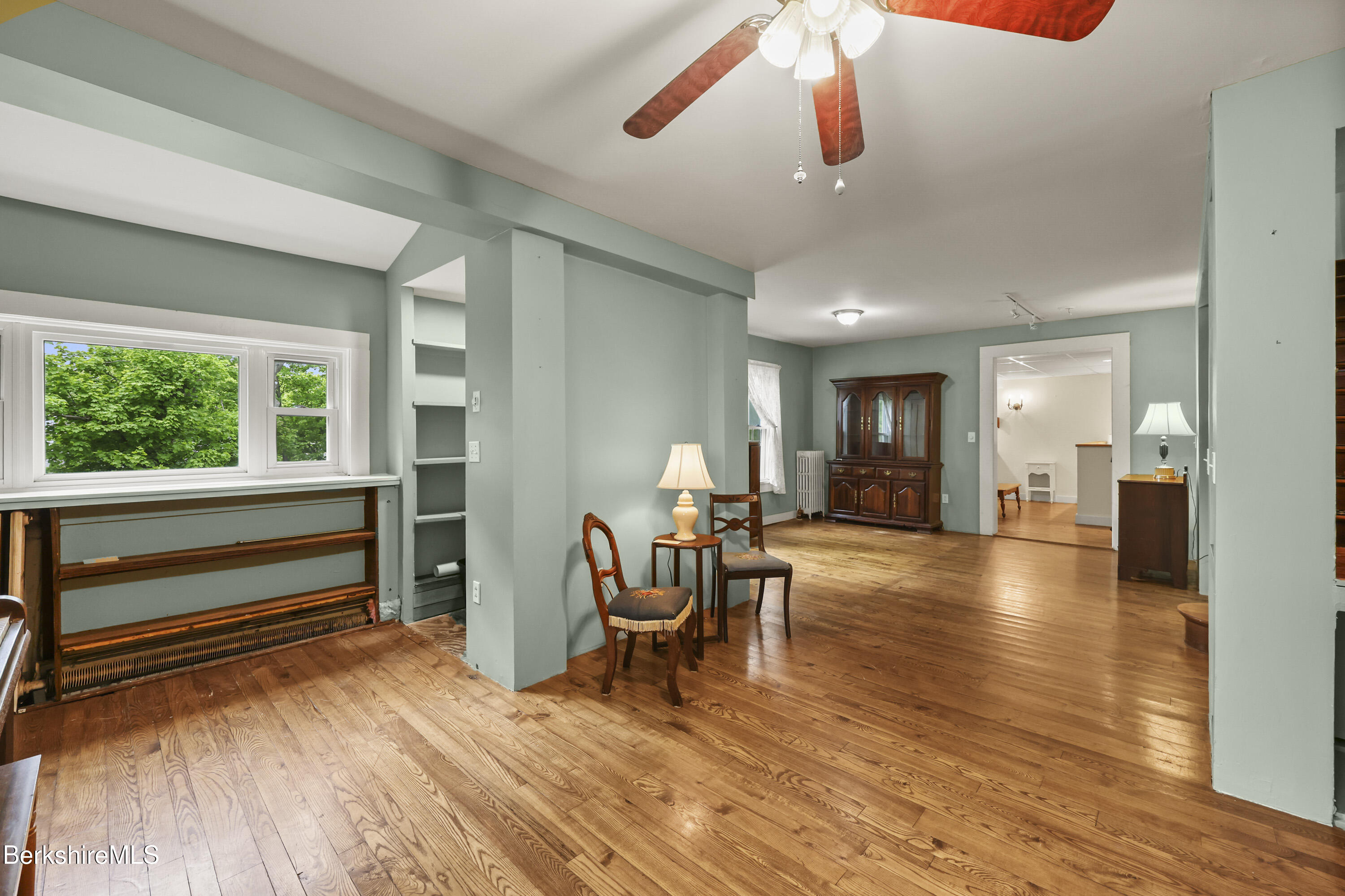 Living room with wide-plank hardwood floors, sage-green walls, built-in shelves, and a large leafy-window view.