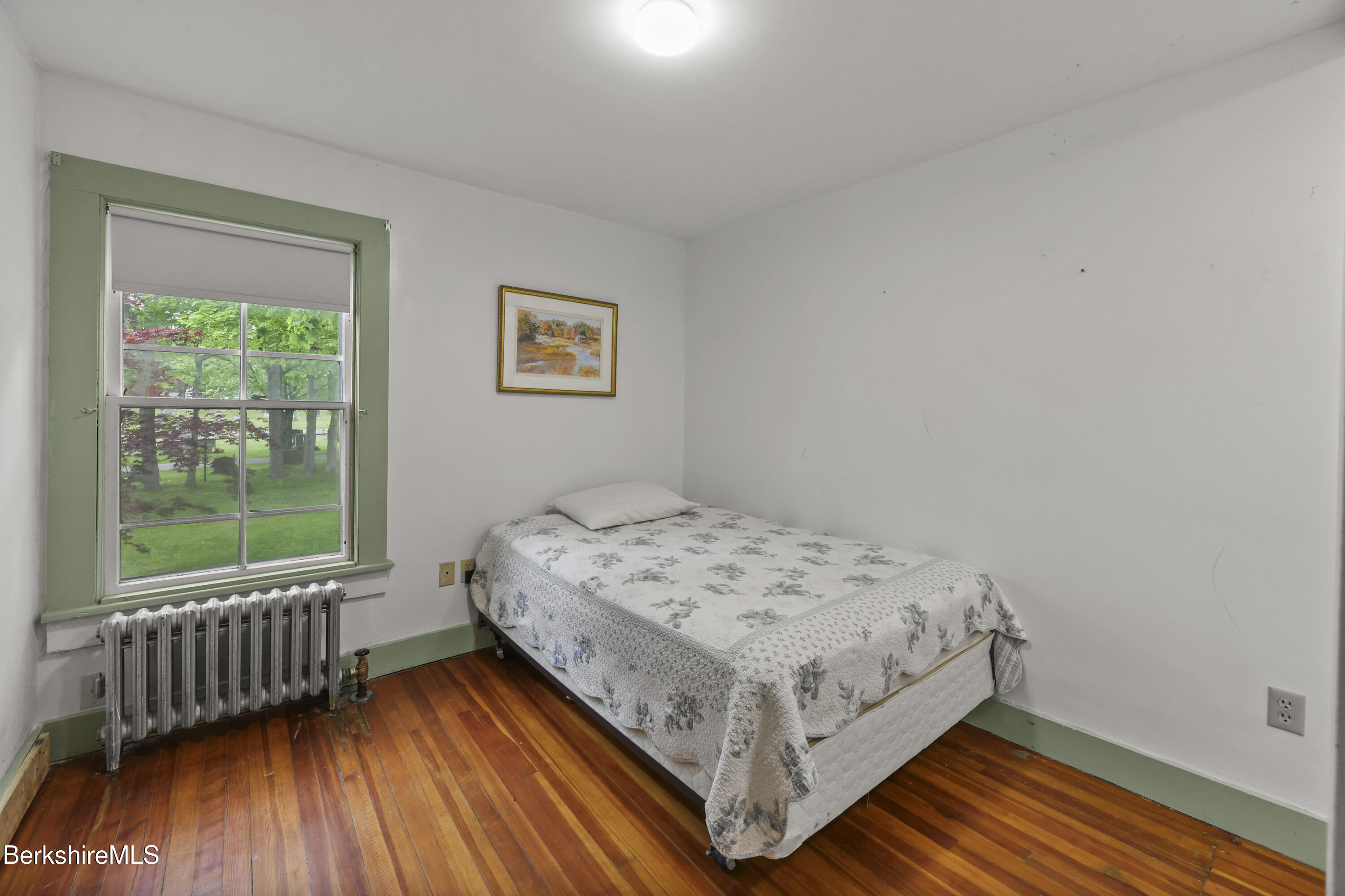 Bedroom with a floral-covered bed, polished wood floors, and a green-trimmed window overlooking trees.