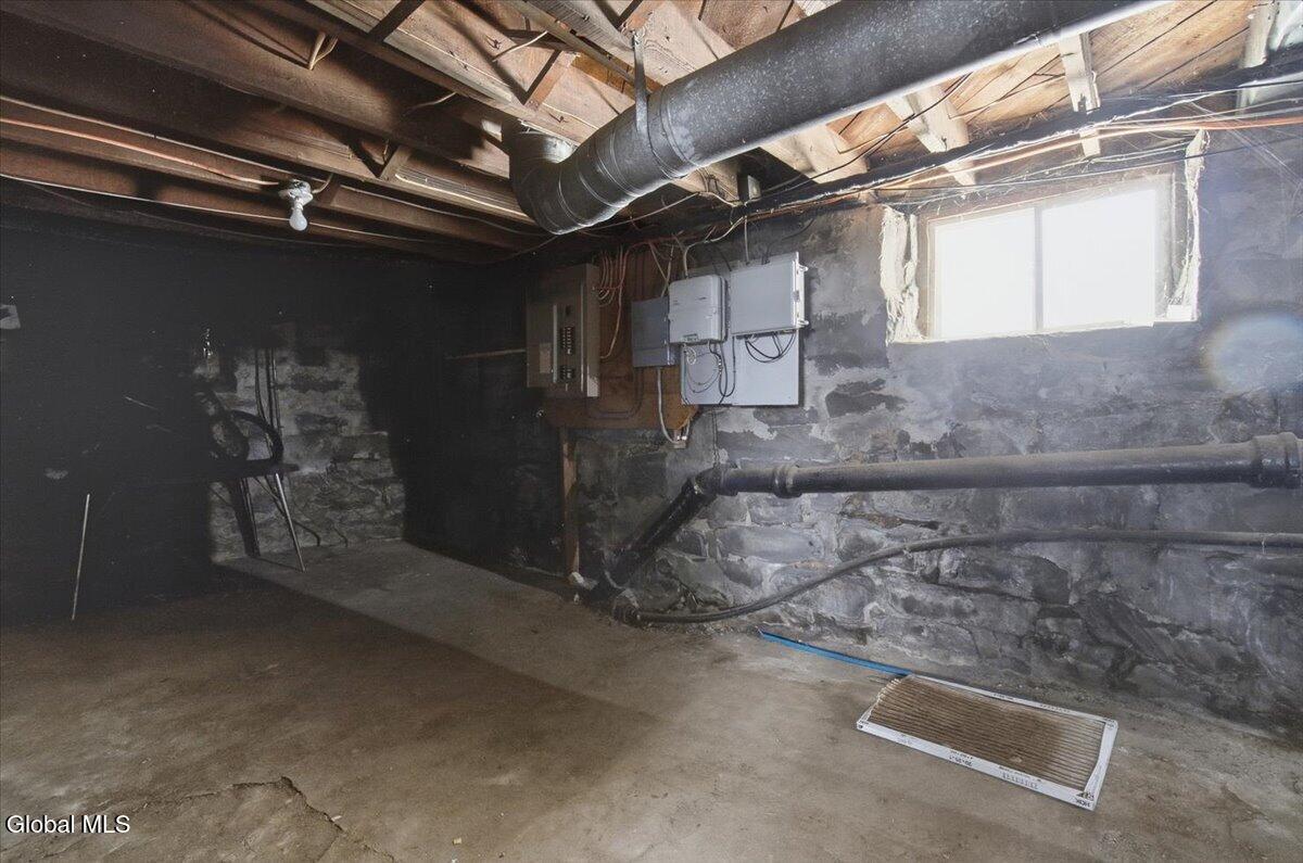 Unfinished basement with exposed stone walls, ceiling joists, utility pipes, and a small window.