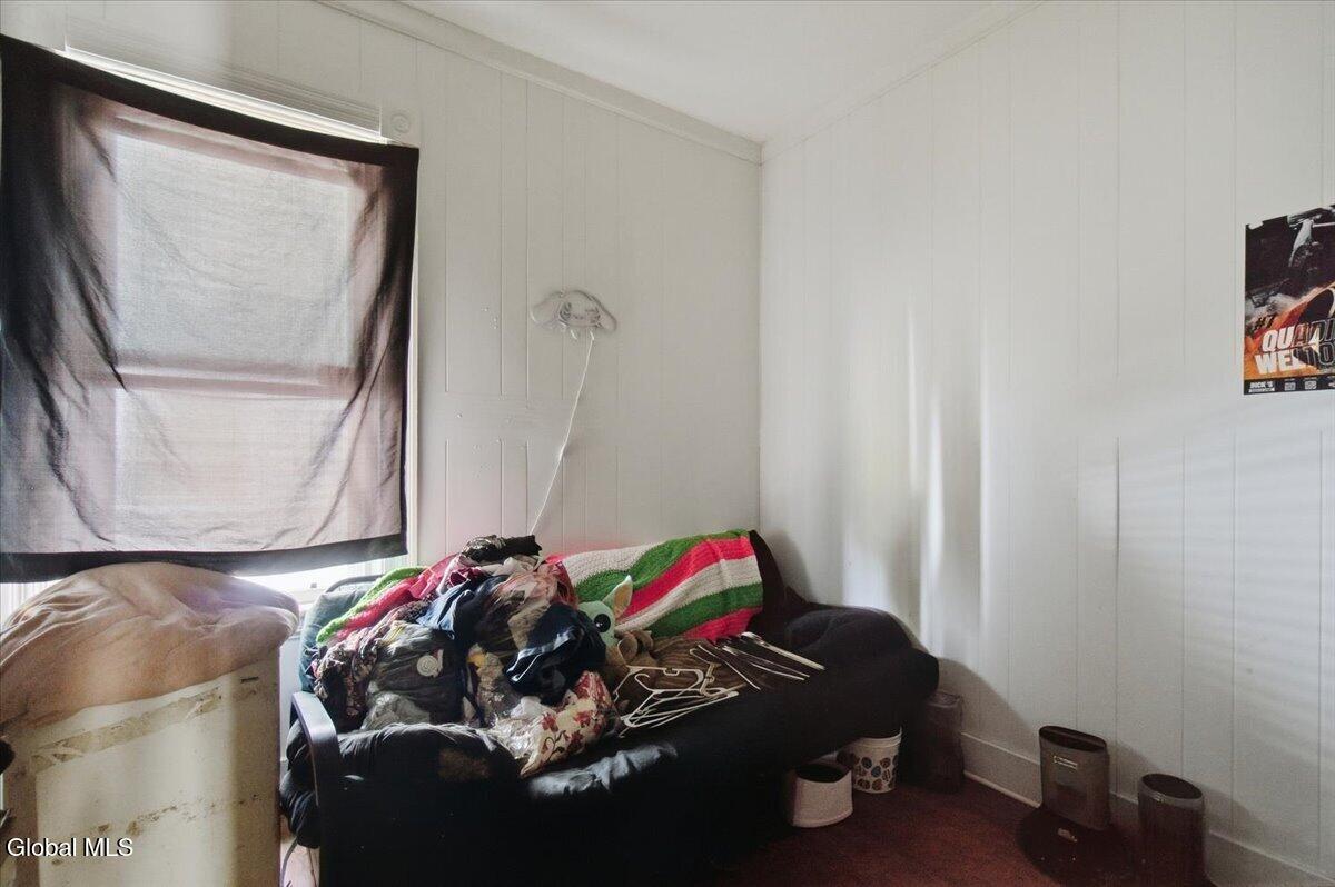 Room with a dark sofa piled with clothes and hangers beneath a curtained window.