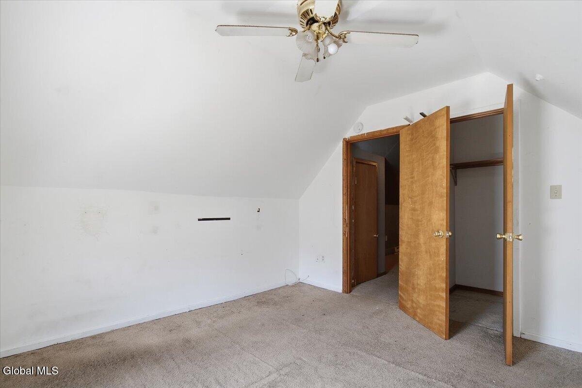 Empty bedroom with beige carpet, sloped ceiling, ceiling fan, and open wooden closet doors.