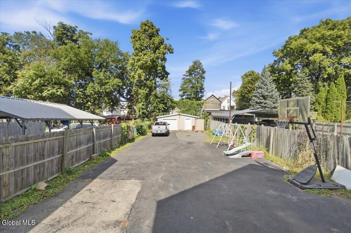 Fenced backyard with a paved driveway, detached garage, basketball hoop, and swings.