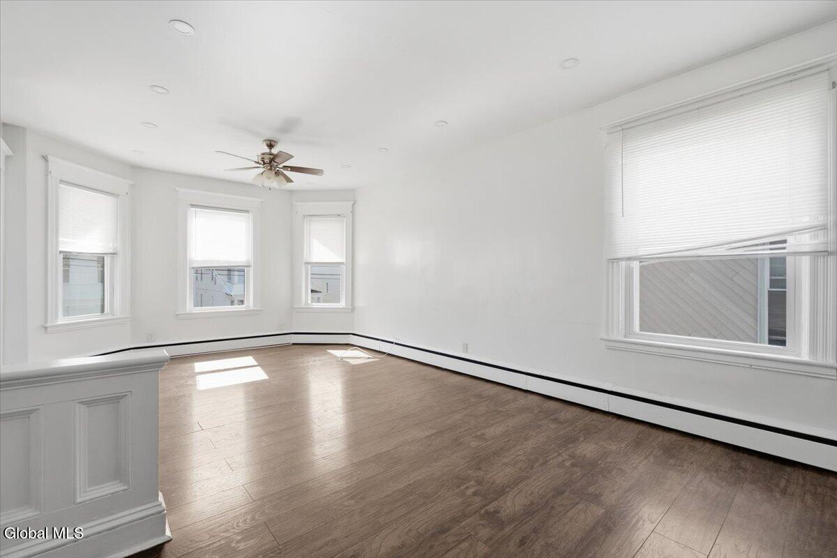 Vacant room with brown wood flooring, white walls, several windows, and a ceiling fan.