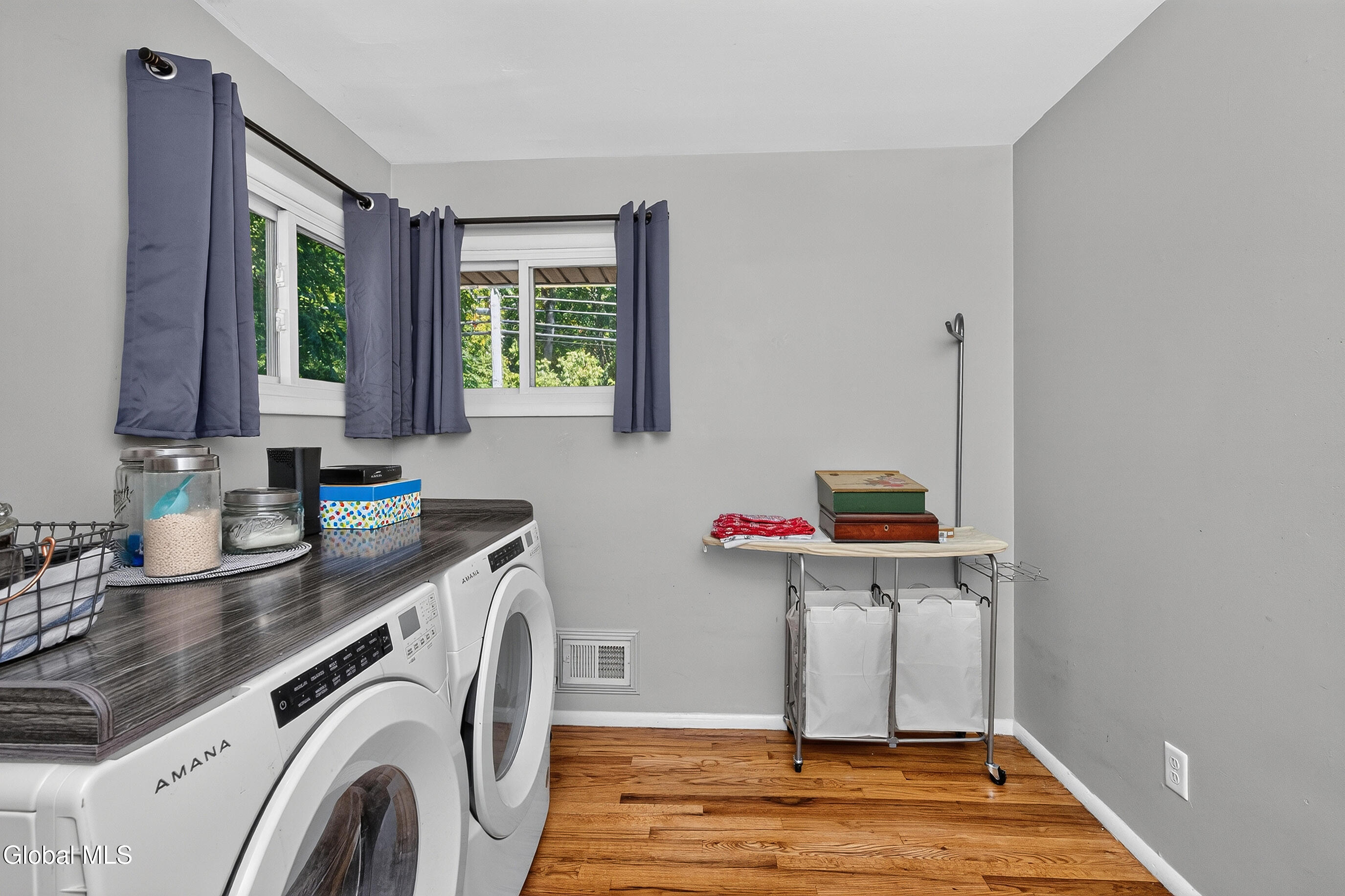 Laundry room with front-loading washer and dryer, curtained windows, and wood flooring.