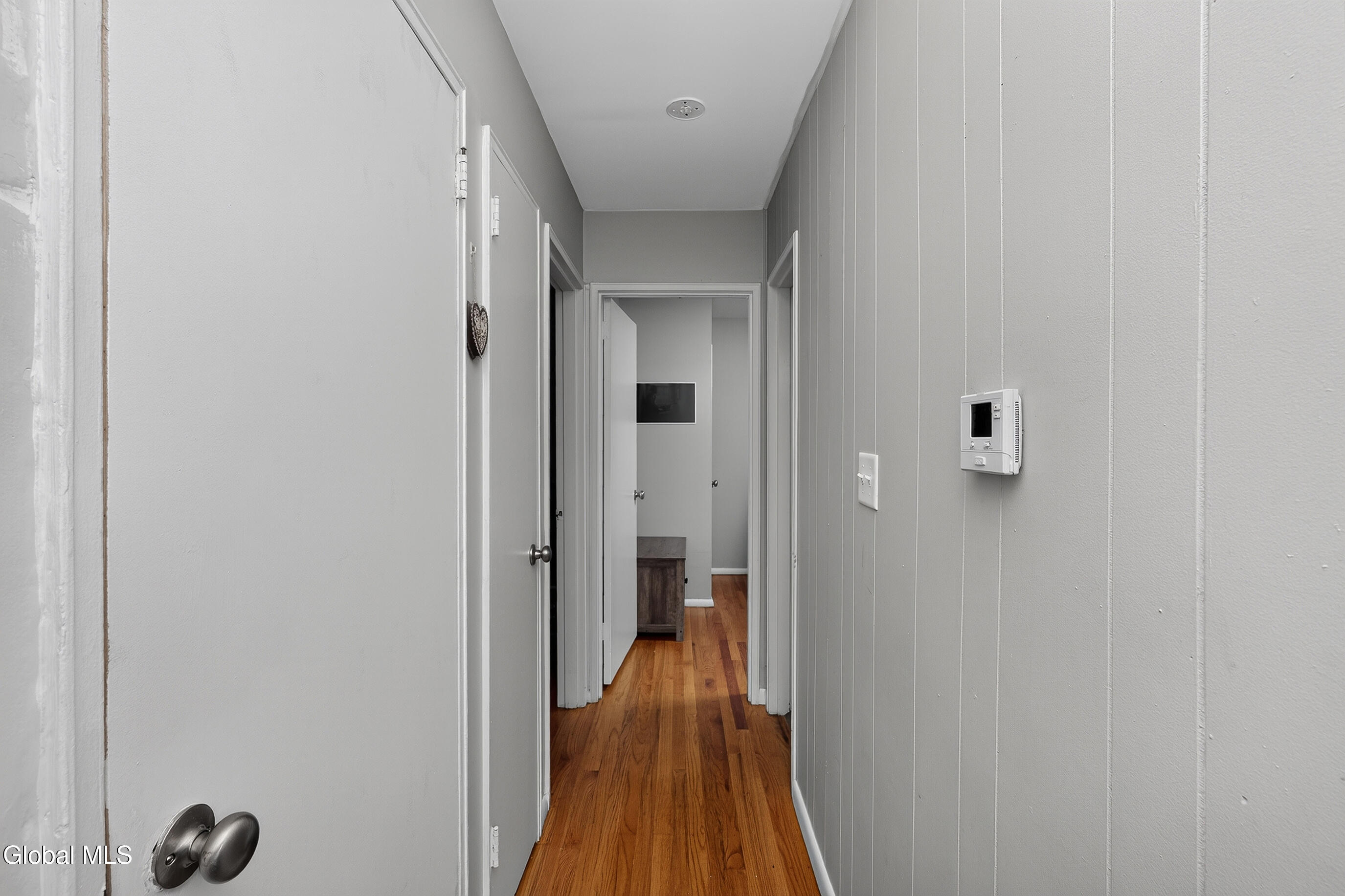 Narrow gray hallway with white doors, hardwood flooring, and a wall-mounted control panel.