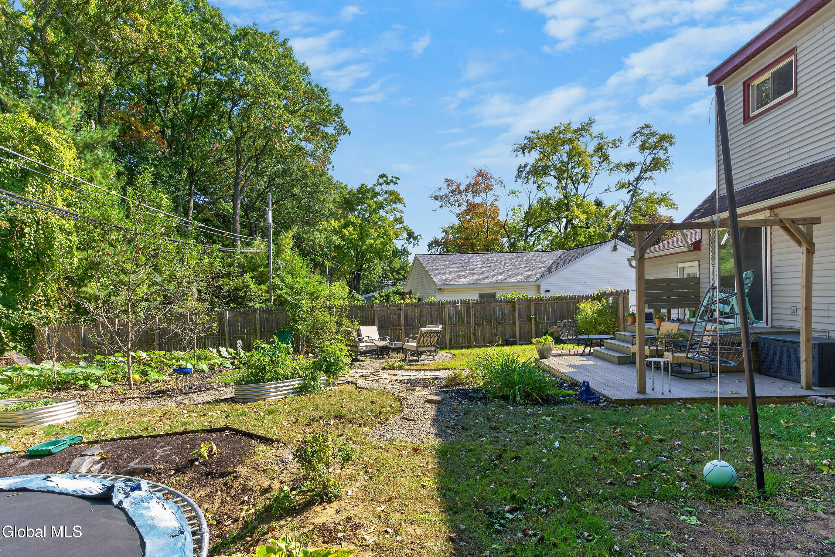 Fenced backyard with a wooden deck, garden beds, outdoor seating, and a trampoline.