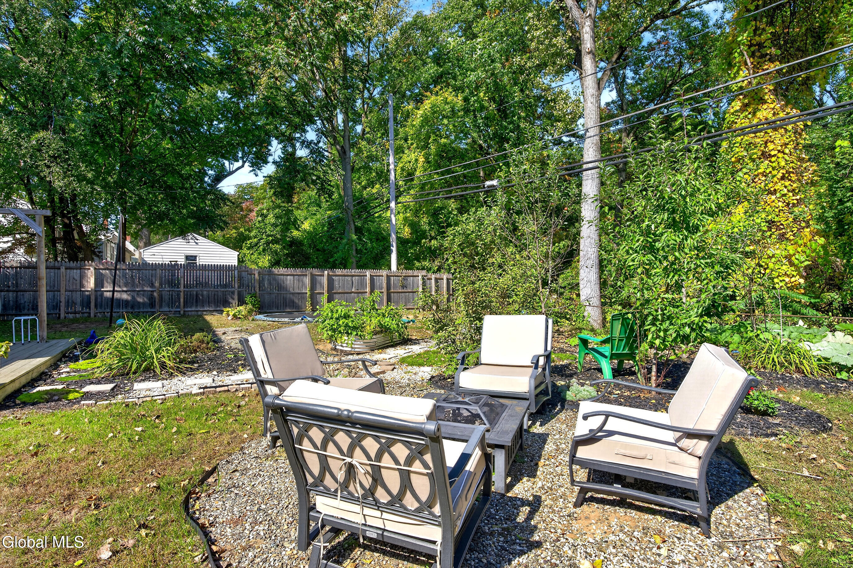 Backyard patio with cushioned seating on gravel, surrounded by garden beds, mature trees, and a wooden fence.