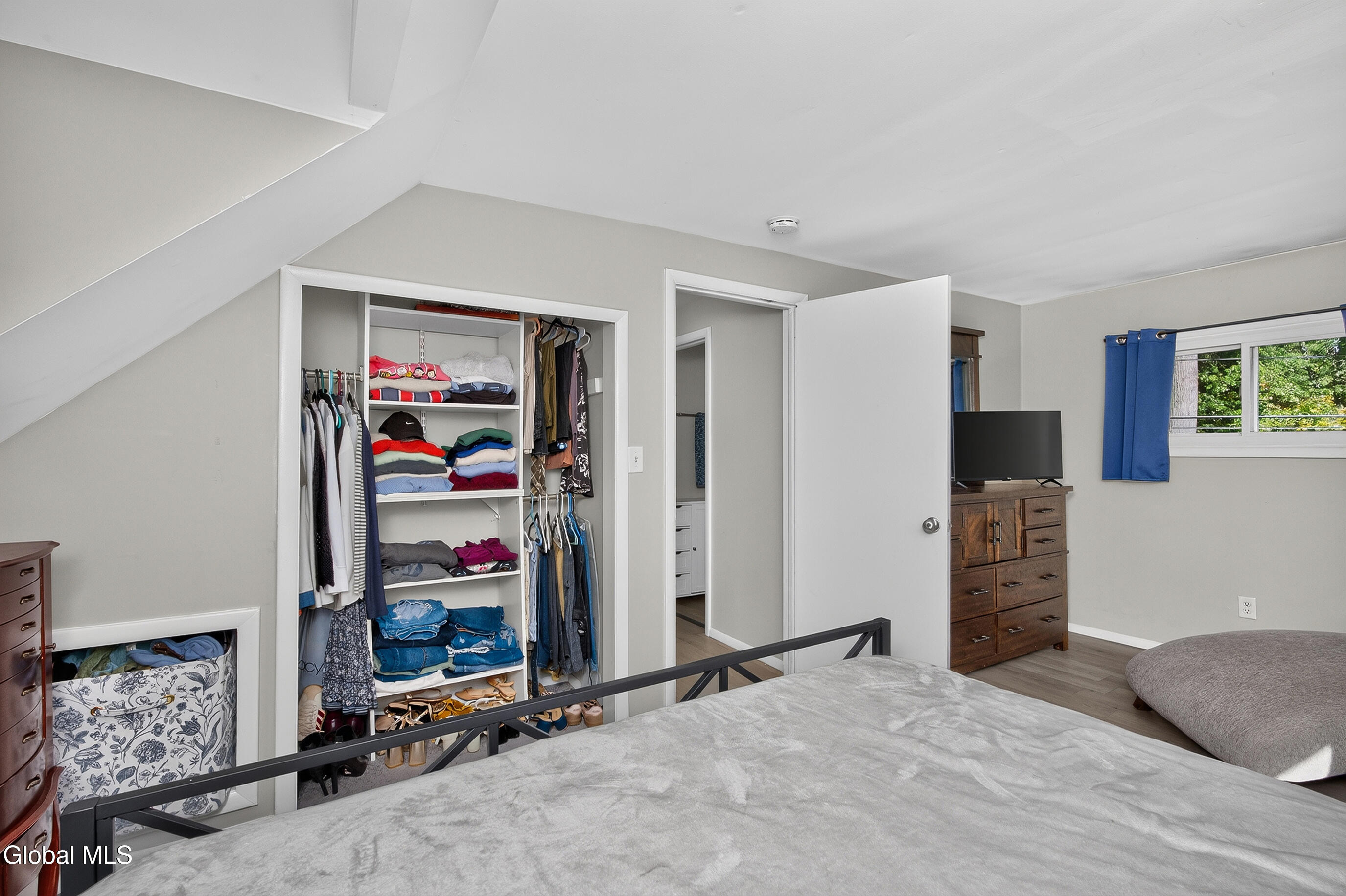 Attic bedroom with a sloped ceiling, open closet, wooden dresser, television, and blue-curtained window.
