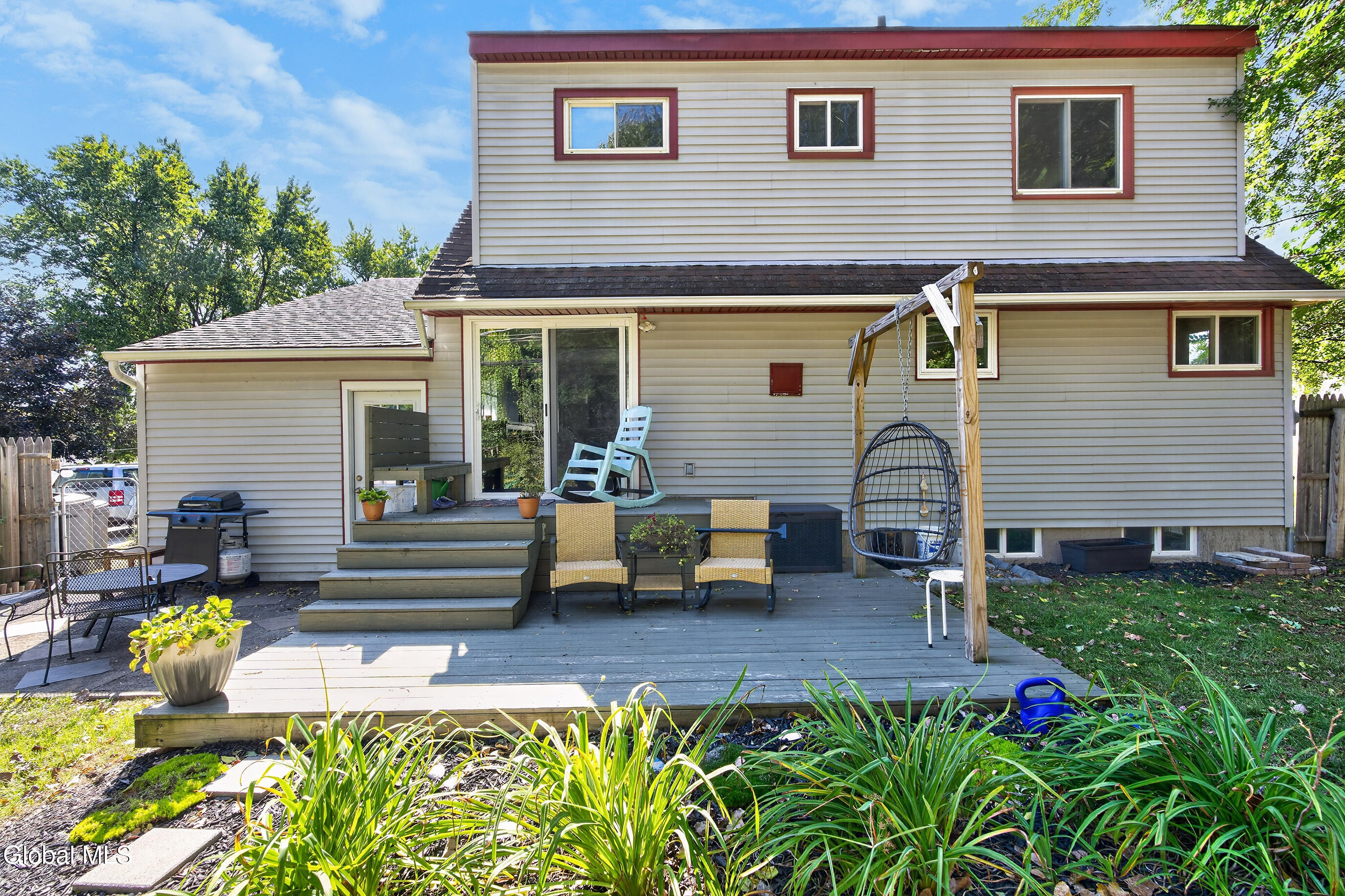 Rear of a beige two-story house with a furnished deck, hanging egg chair, and garden beds.