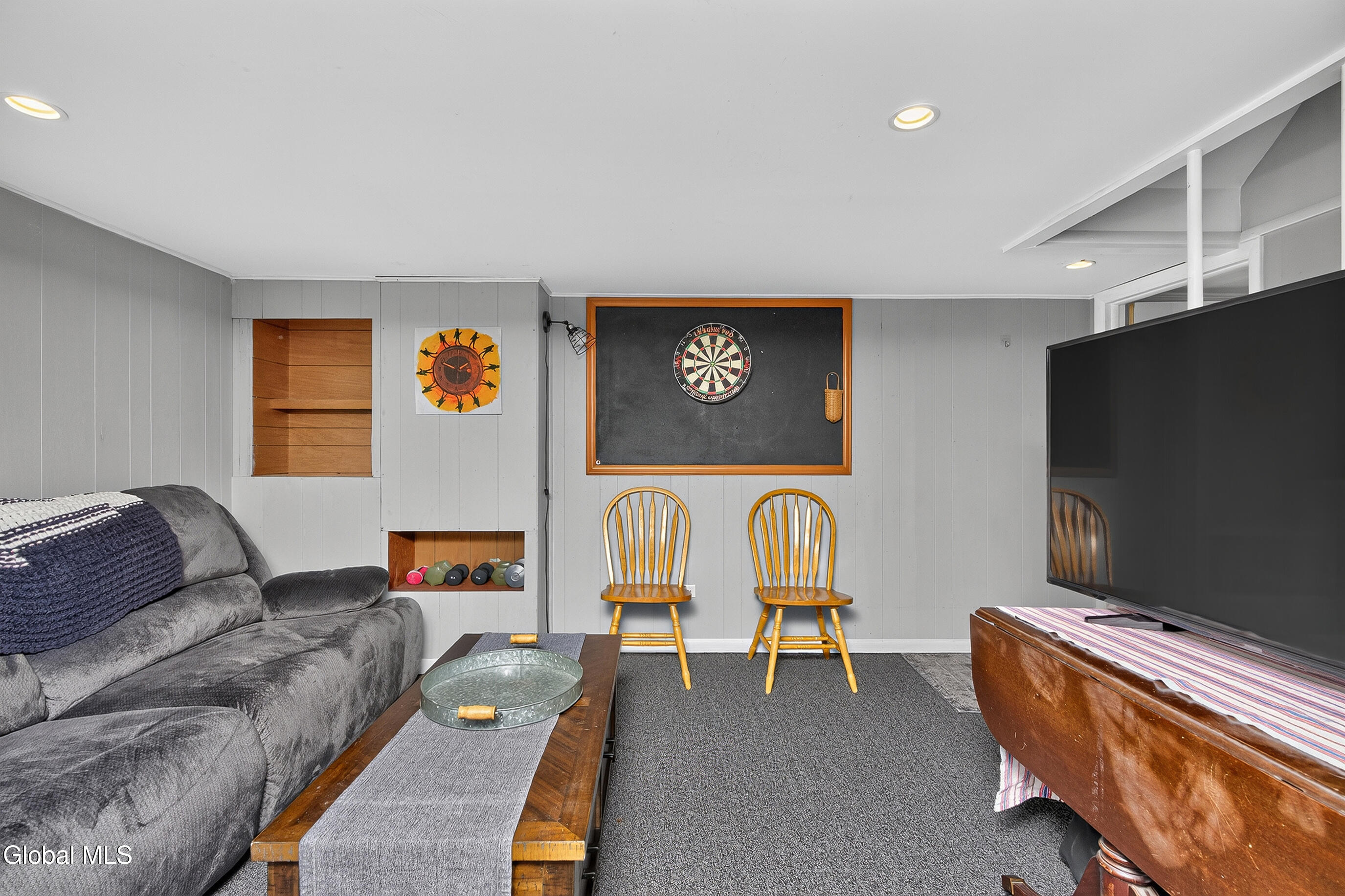 Basement recreation room with a gray sectional, dartboard, wooden chairs, and large television.