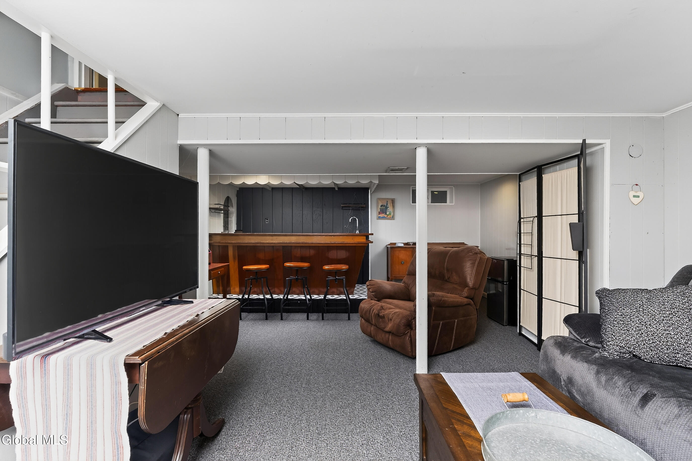 Basement recreation room with a wooden bar, three stools, recliners, and a large television.
