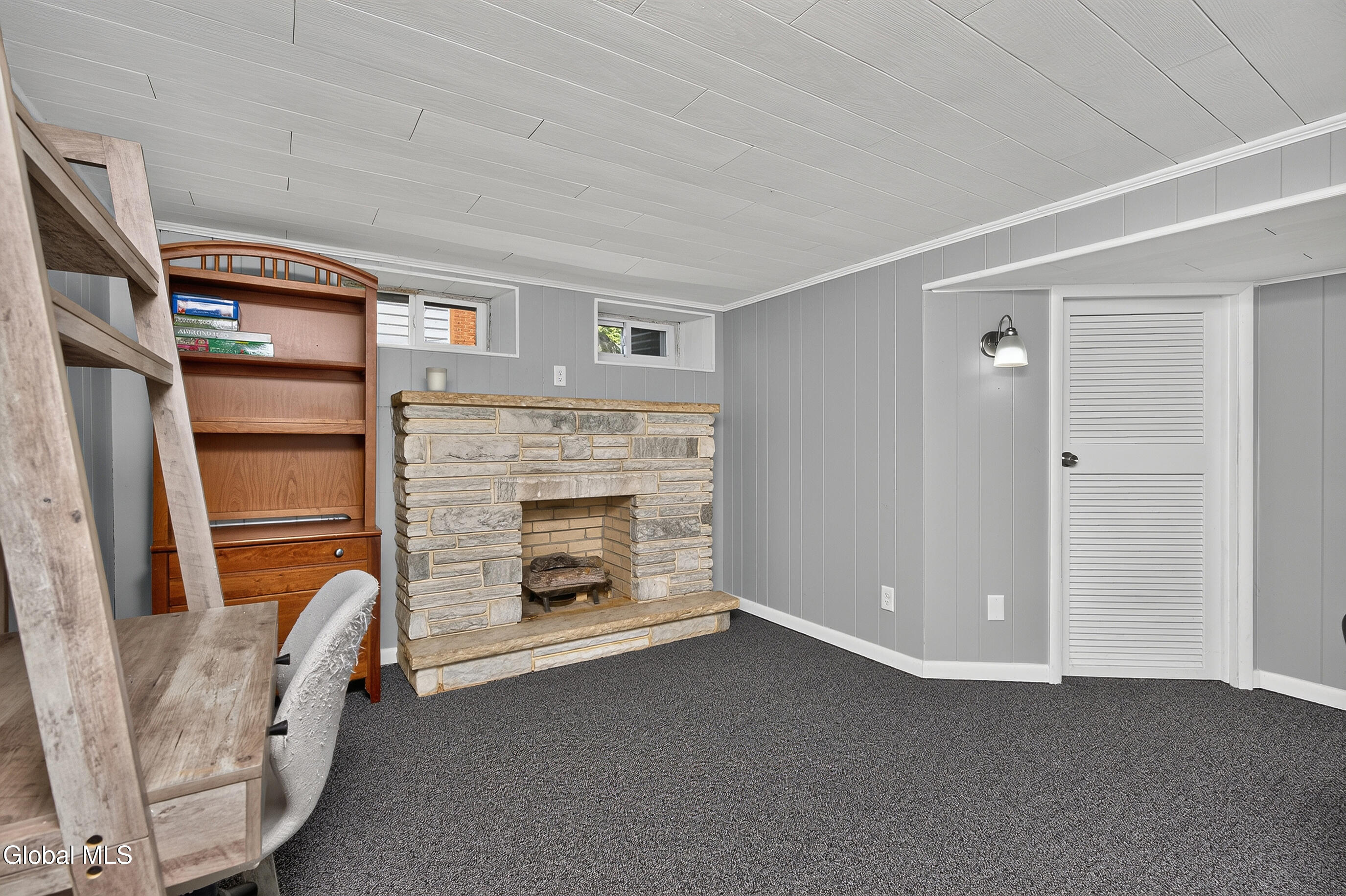 Finished basement with a stone fireplace, built-in wooden shelving, and gray paneled walls.