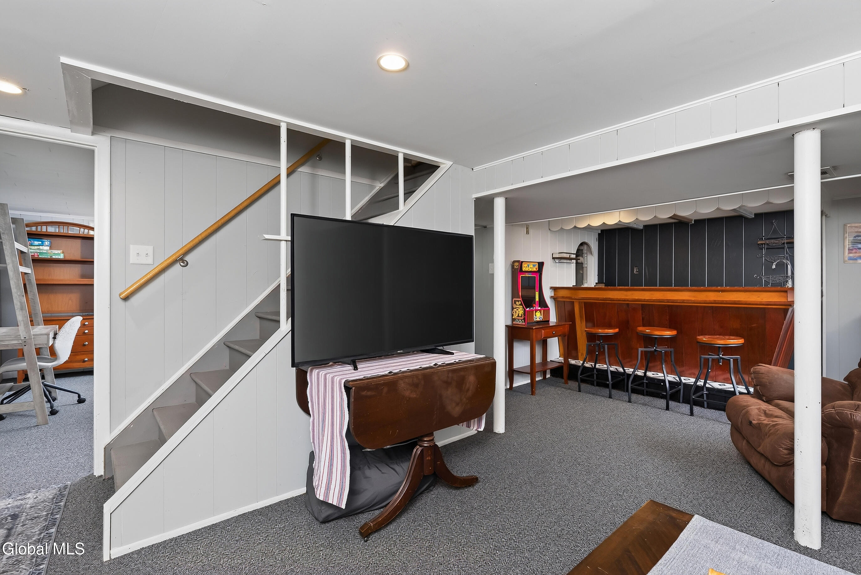 Basement recreation room with a wooden bar, stools, staircase, and large television.