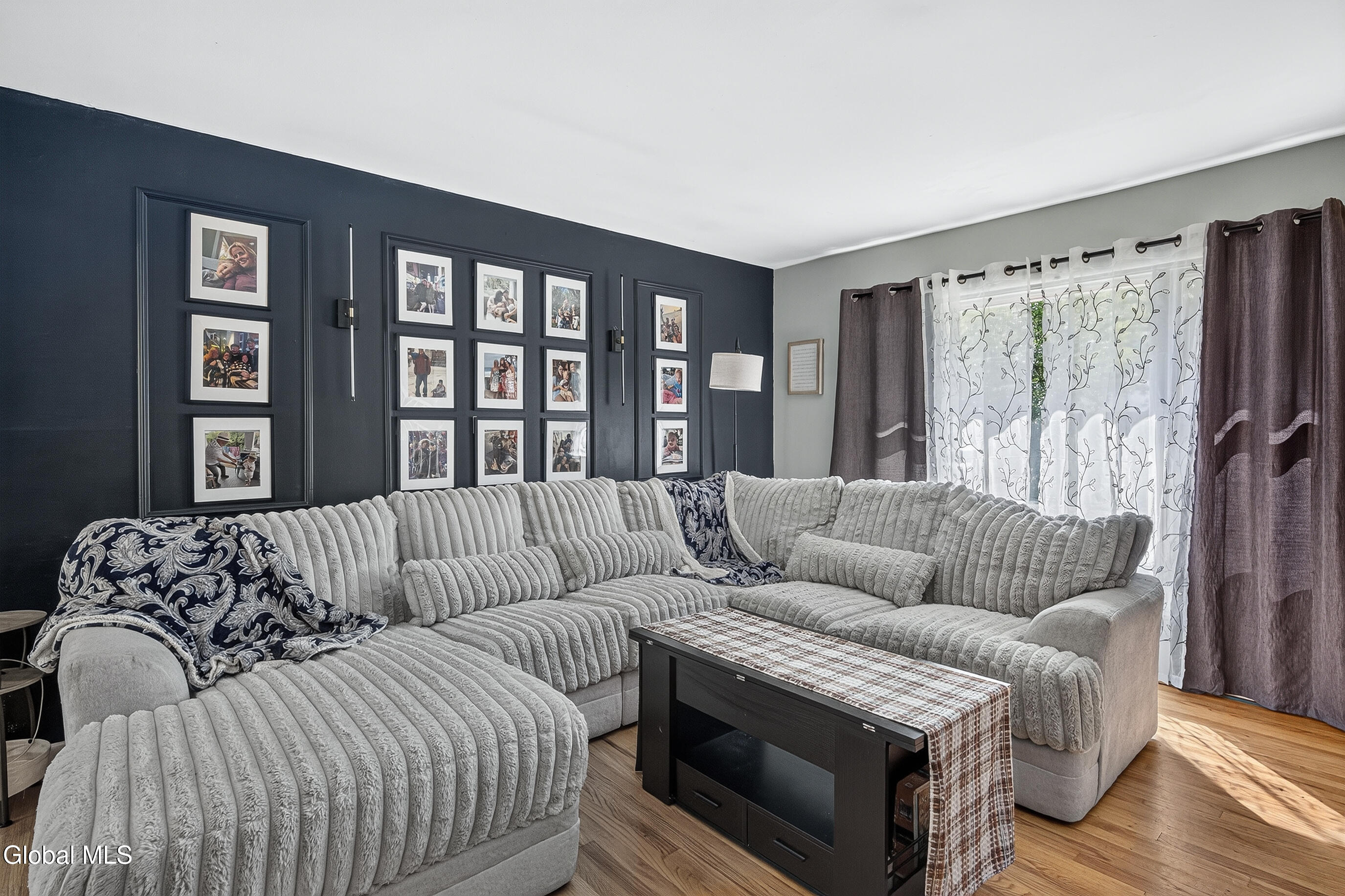 Living room with a light-gray sectional, navy gallery wall, and patterned curtains over a bright window.