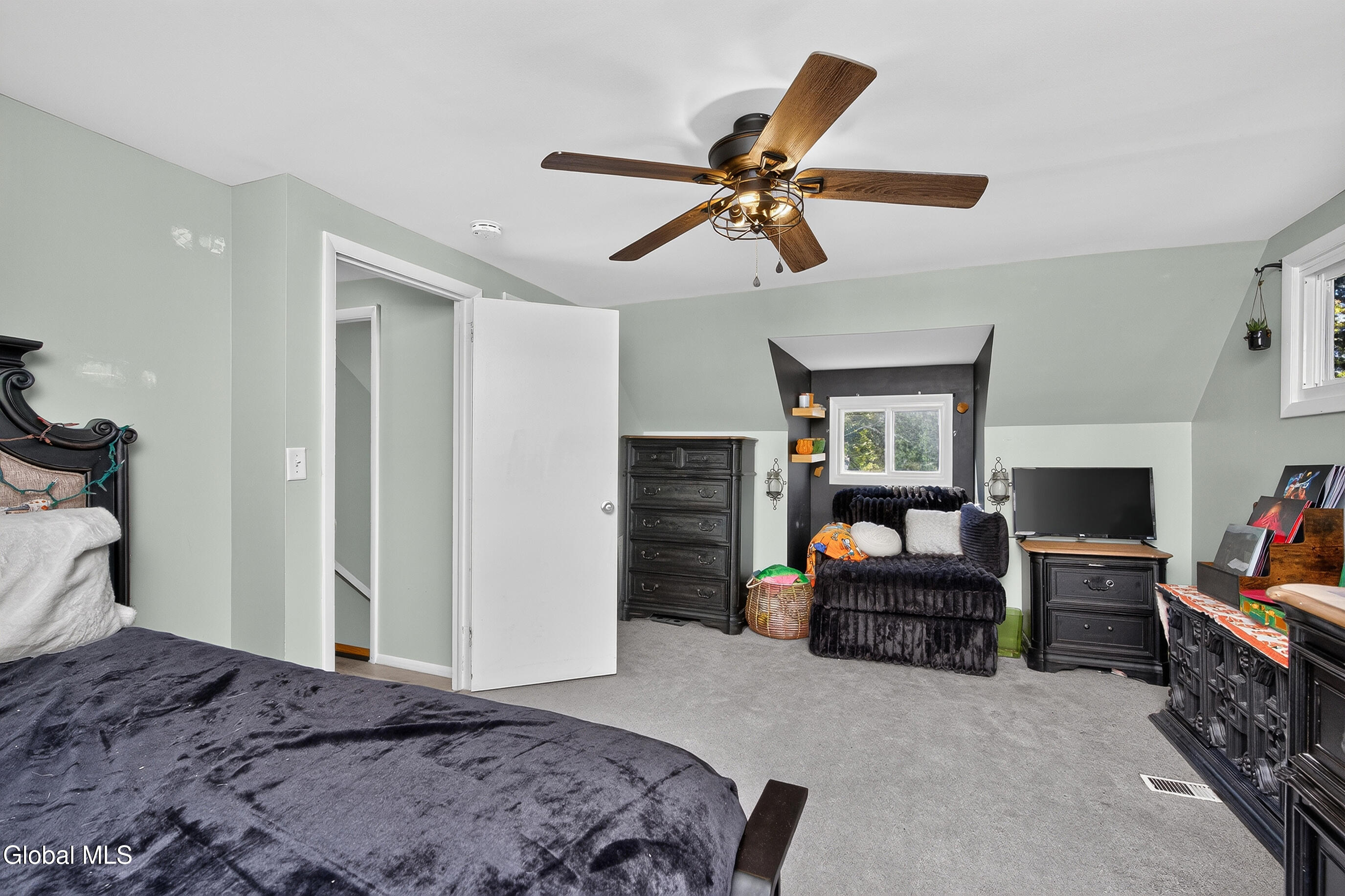 Bedroom with a dark bed, wooden-blade ceiling fan, and loveseat beneath a dormer window.