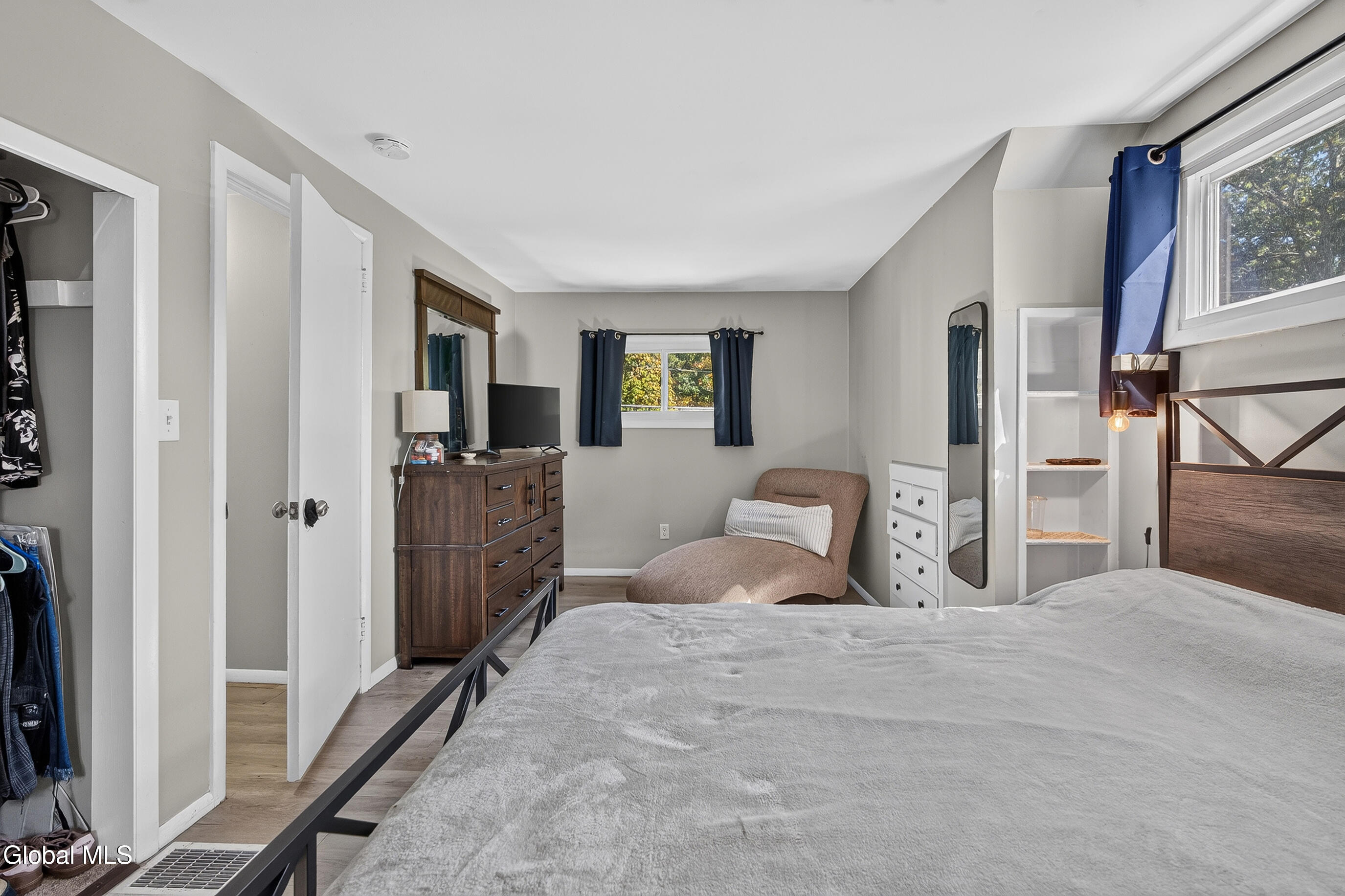 Furnished bedroom with a large gray bed, dark wood dresser, chaise lounge, and open closet.