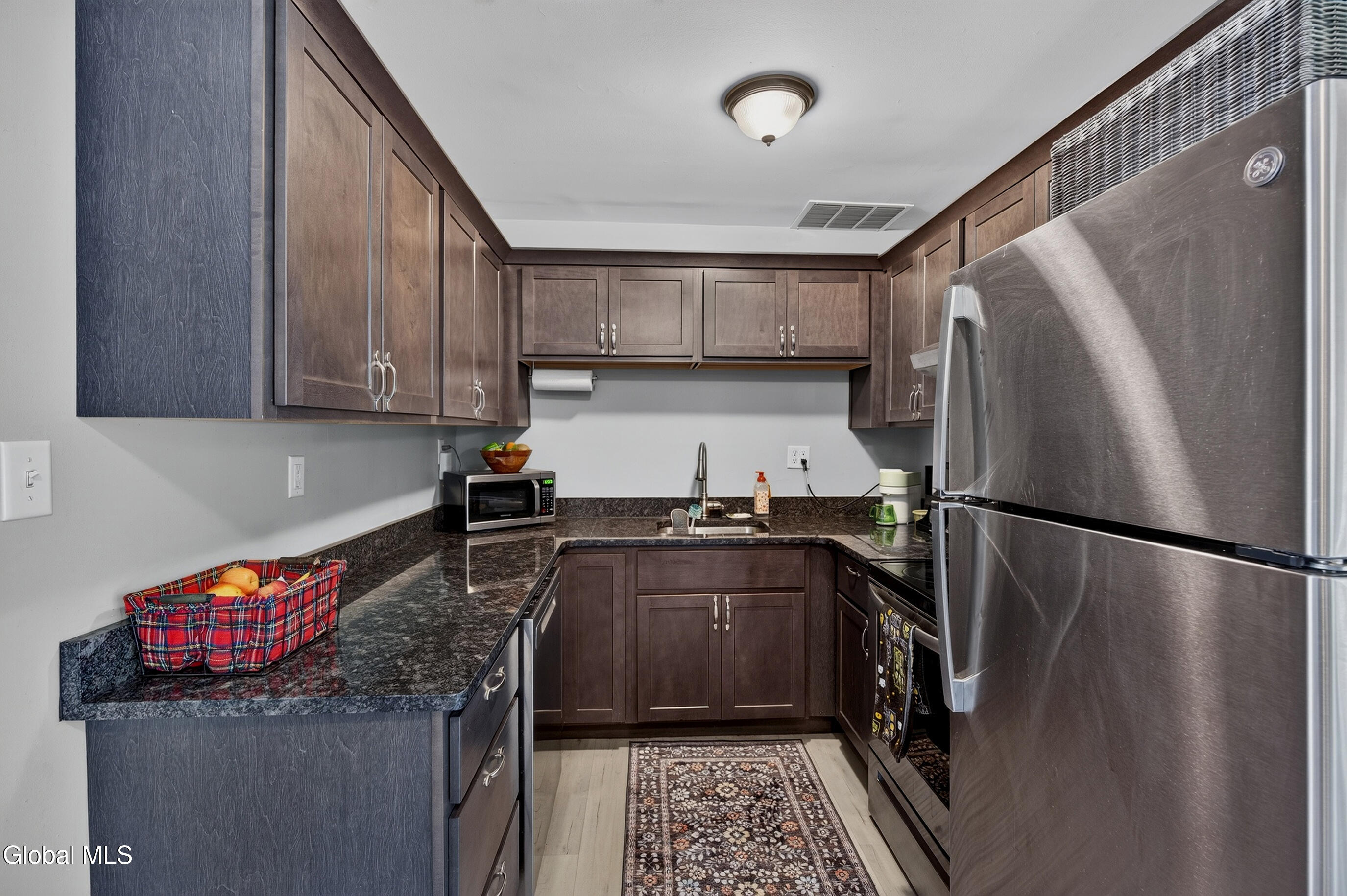 Compact kitchen with dark wood cabinets, granite counters, stainless steel appliances, and a patterned runner.