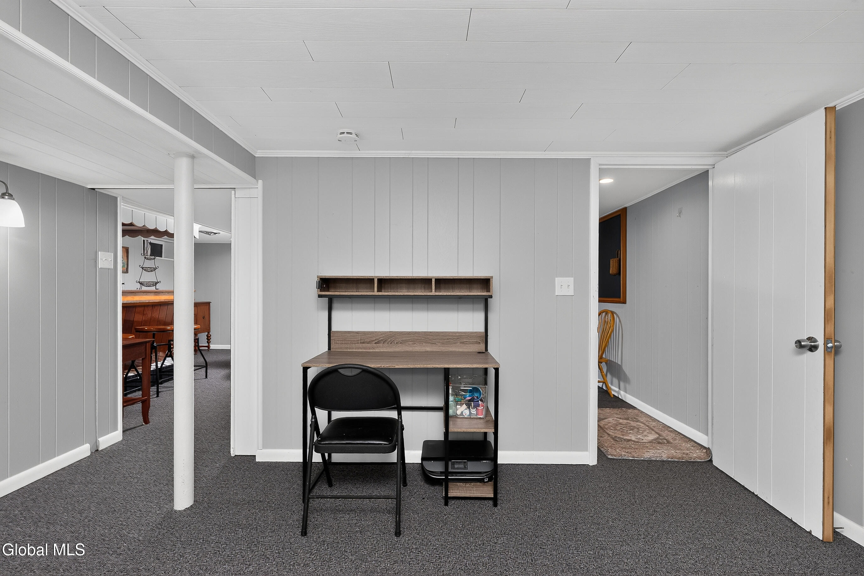Basement office with a desk and shelving, gray paneled walls, and dark carpet.