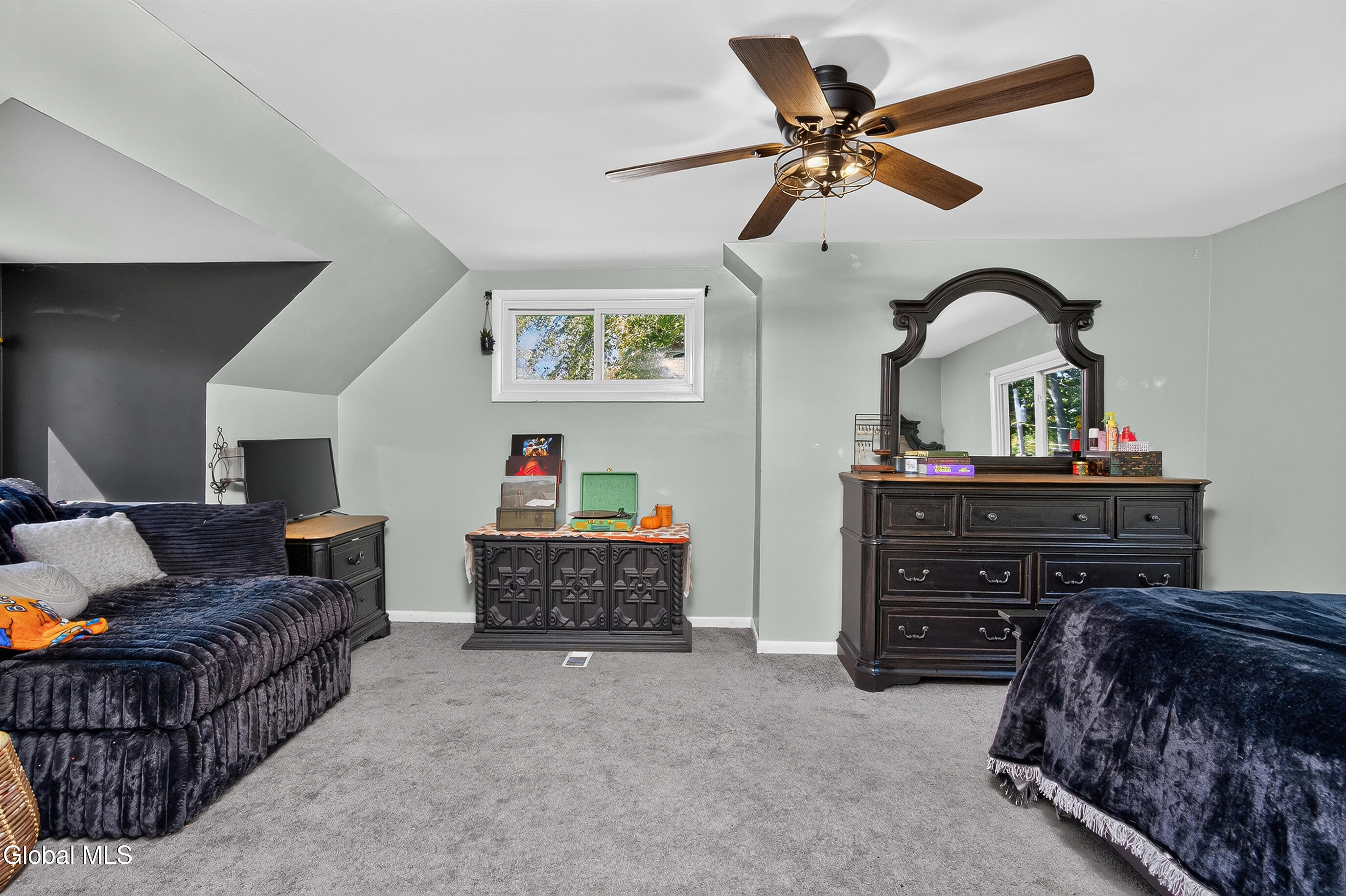 Furnished bedroom with a dark wooden dresser and ornate mirror, ceiling fan, and high window.