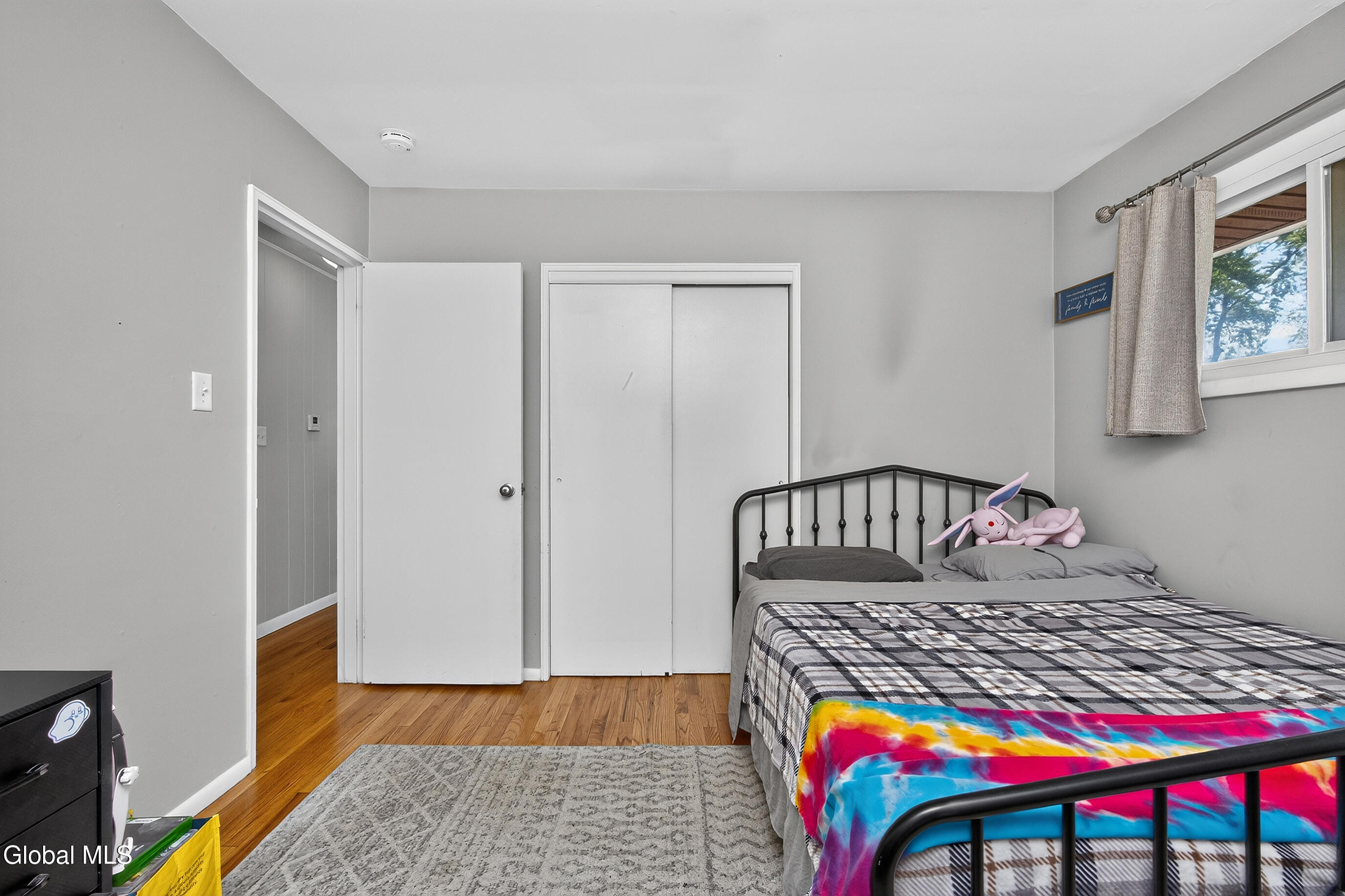 Bedroom with a black metal bed, colorful blanket, hardwood floors, and curtained window.