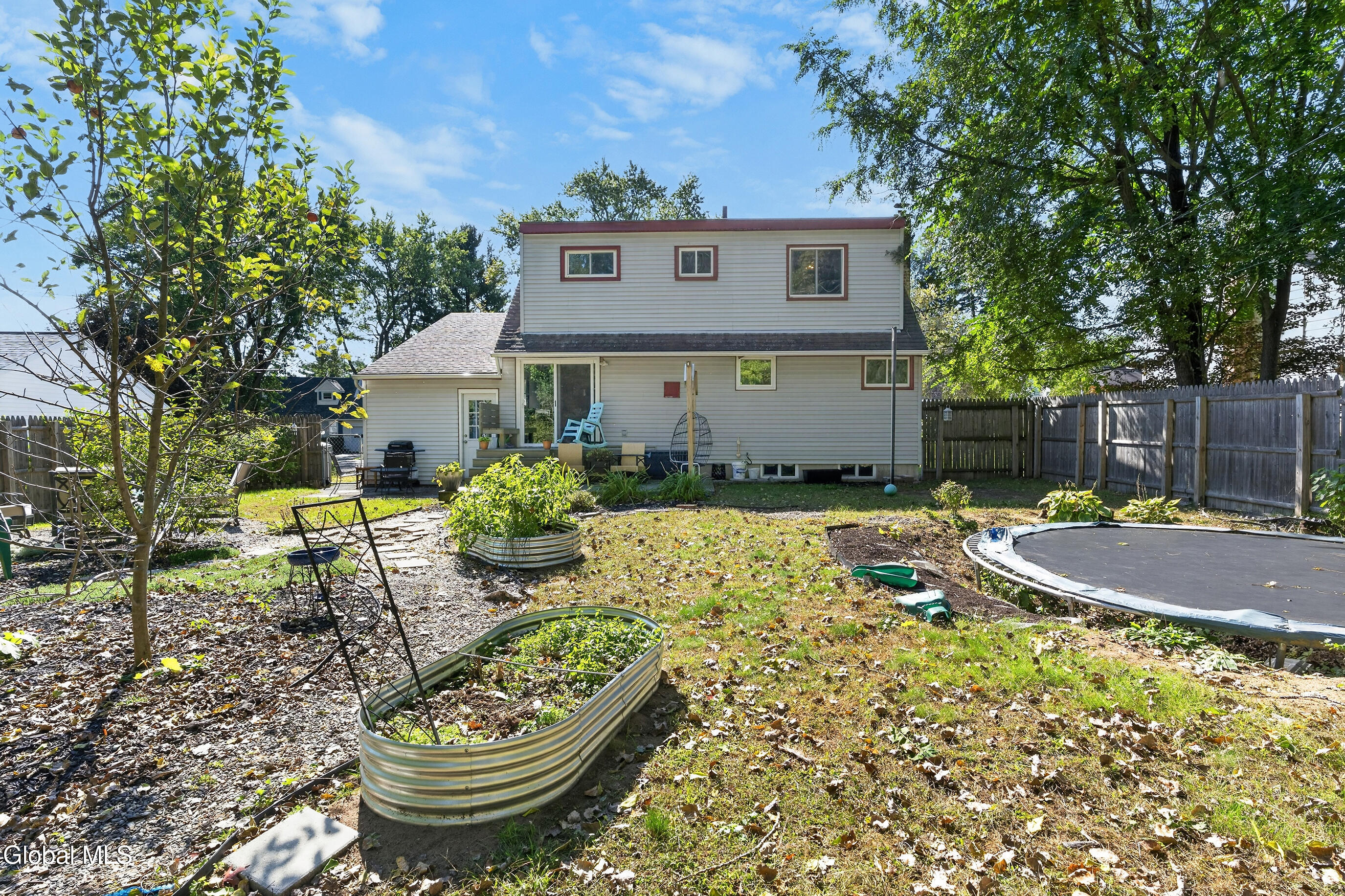 Rear of a two-story house with a fenced yard, raised garden beds, patio seating, and a trampoline.
