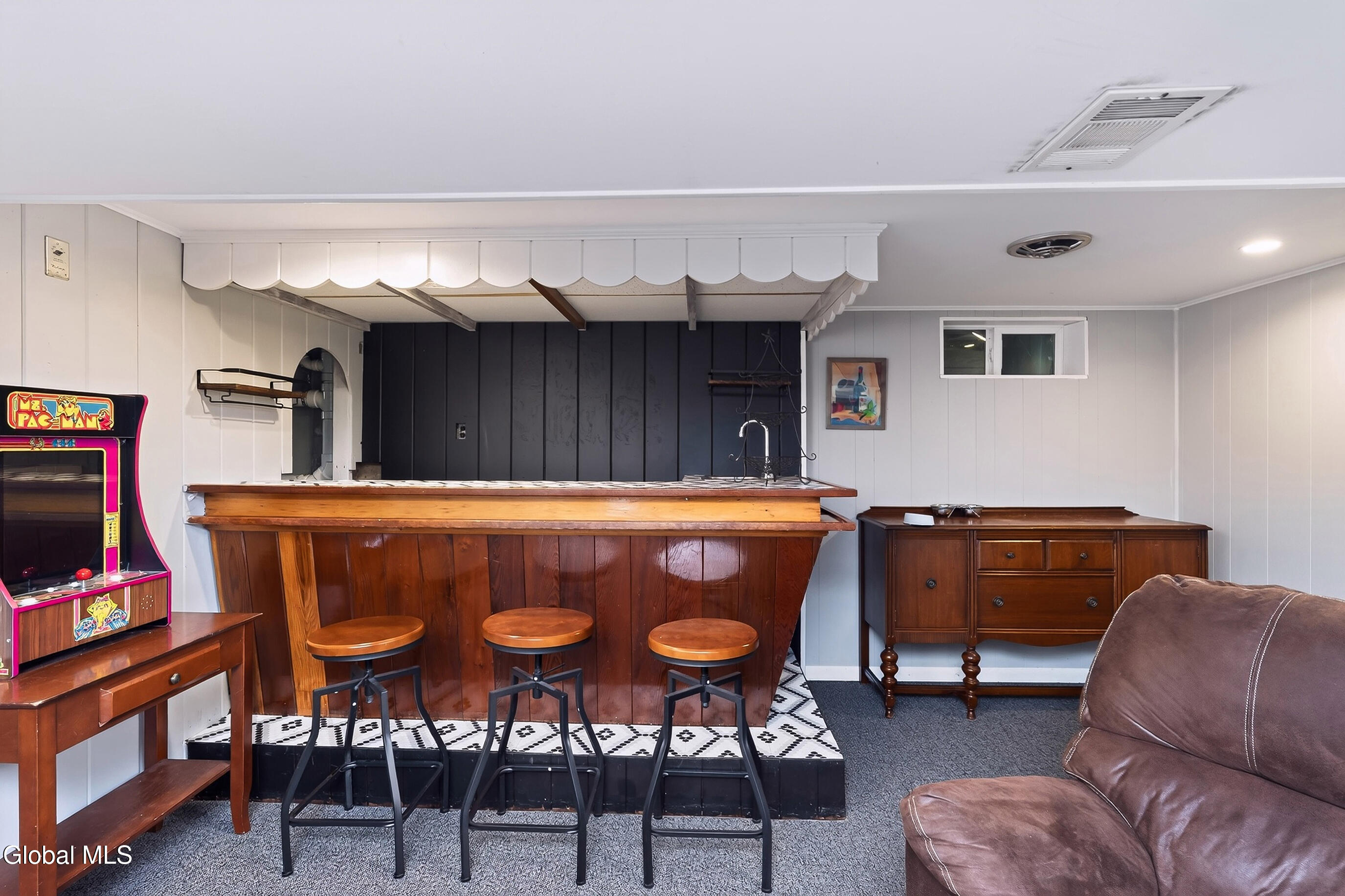 Recreation room with a wooden bar, three stools, a Ms. Pac-Man arcade cabinet, and leather sofa.