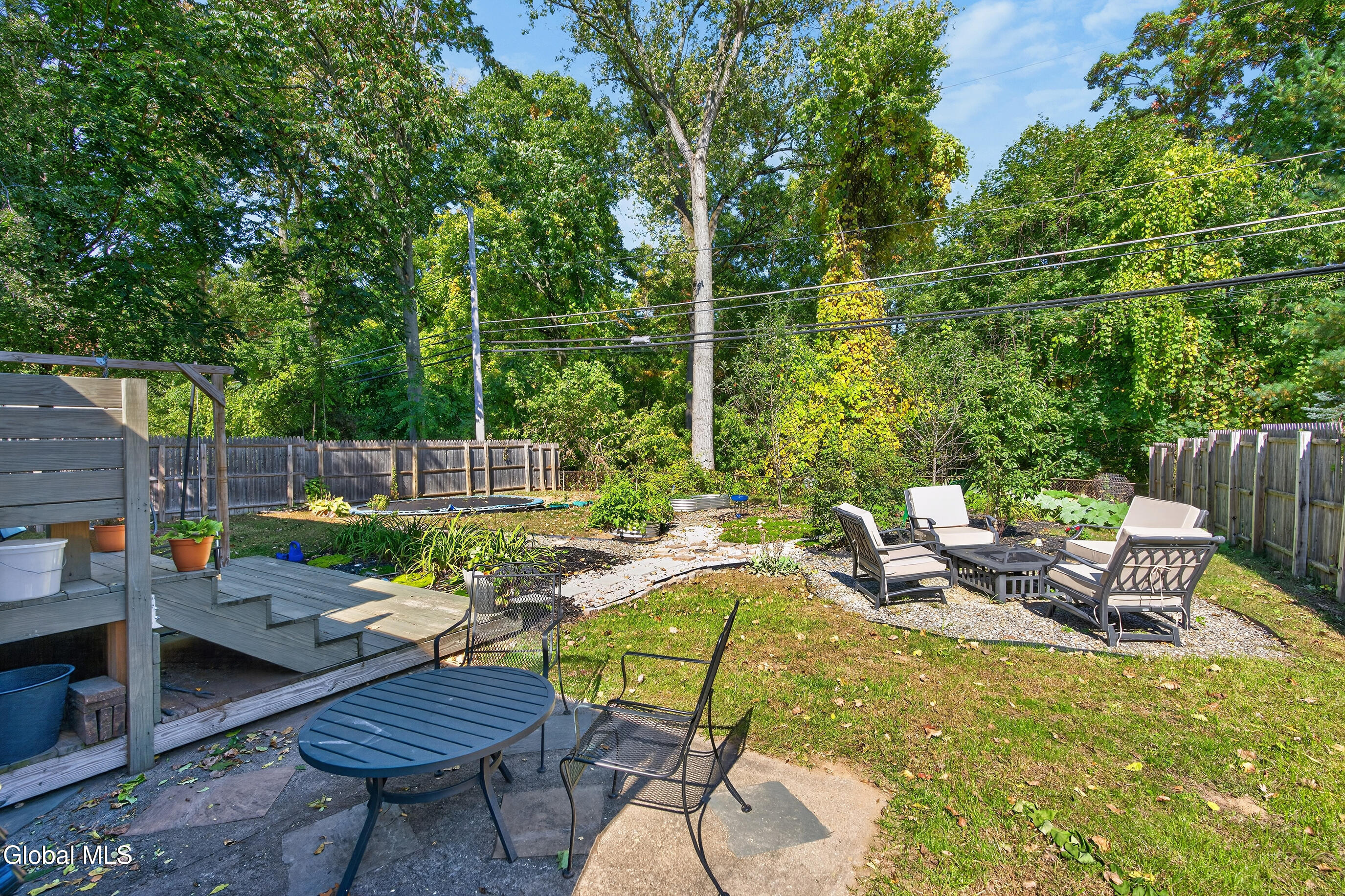 Fenced backyard with a raised deck, garden beds, and a stone patio with outdoor seating.