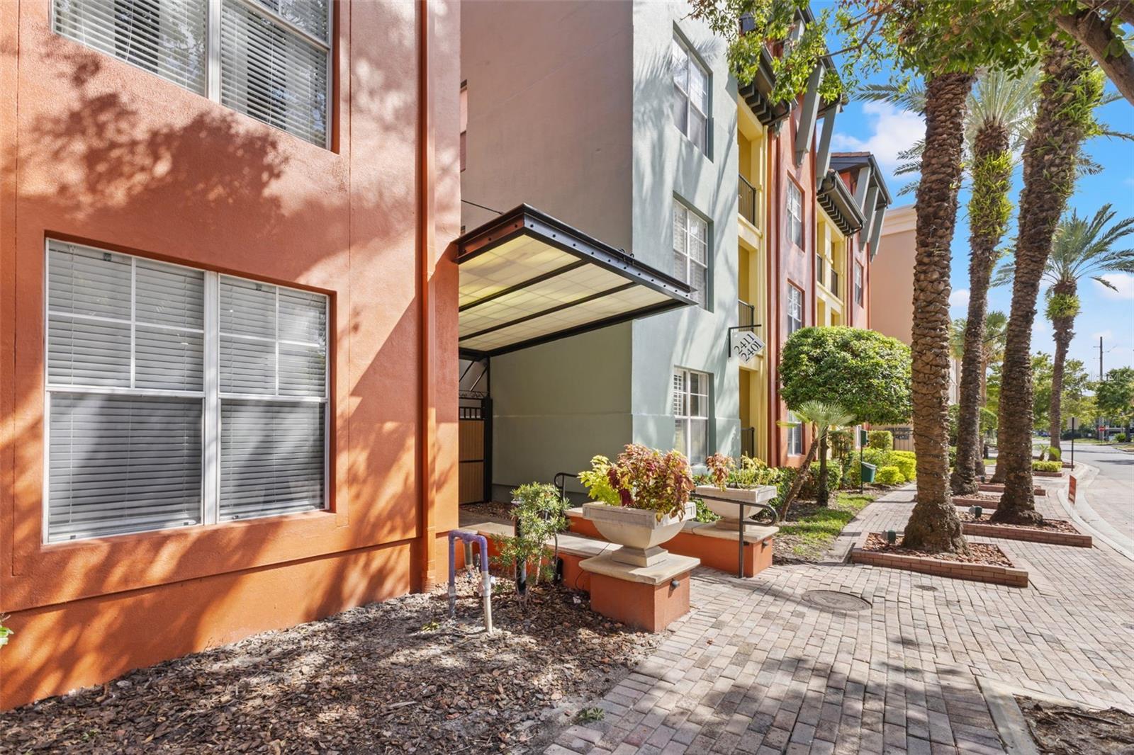 Colorful multistory building entrance with a covered canopy, planters, and palm-lined sidewalk.