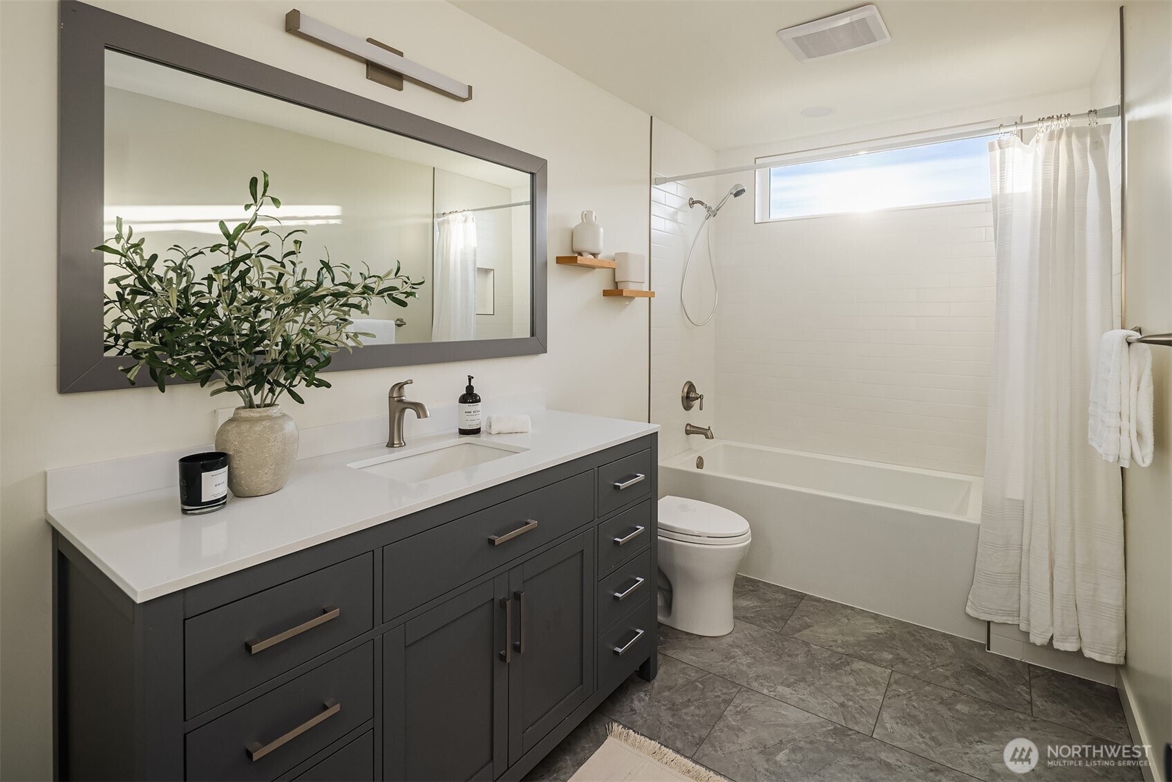 Bathroom with a dark gray vanity, large mirror, and white tub beneath a high window.