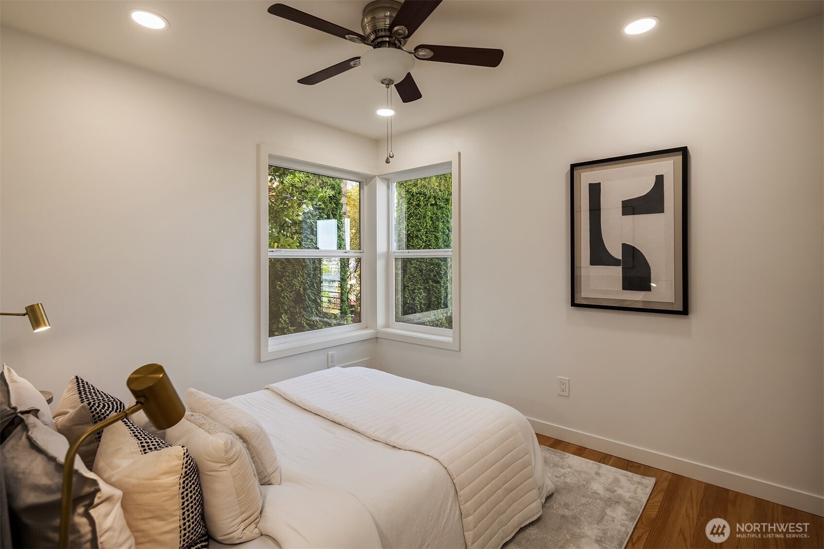 Bedroom with a white bed, corner windows overlooking greenery, and a ceiling fan.