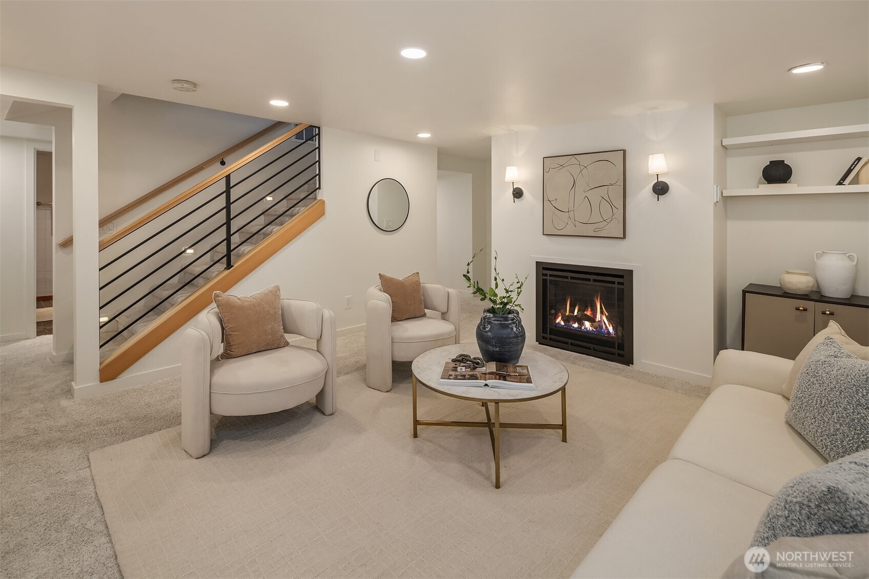 Staged living room with a lit fireplace and staircase with black metal railing.