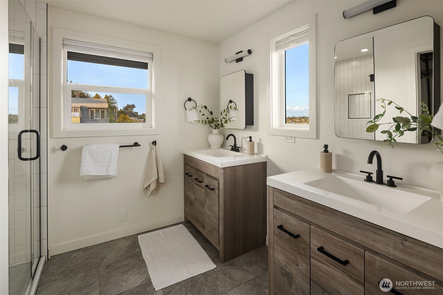 Bright bathroom with two wood vanities, black fixtures, and a glass-enclosed tiled shower.