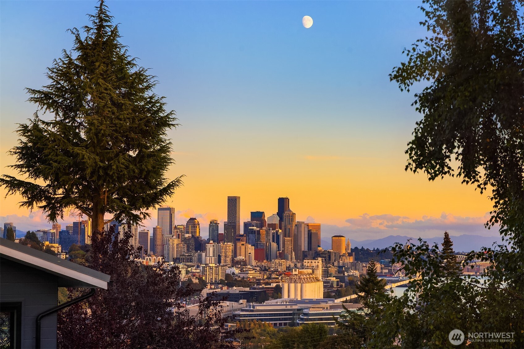 Seattle skyline at sunset framed by trees, with the moon above downtown.