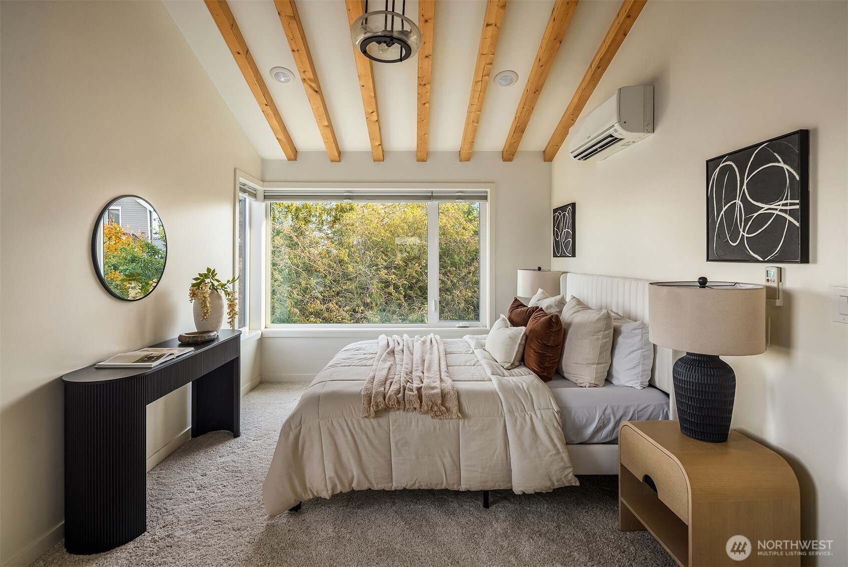 Bedroom with exposed wood ceiling beams and a large window overlooking leafy trees.