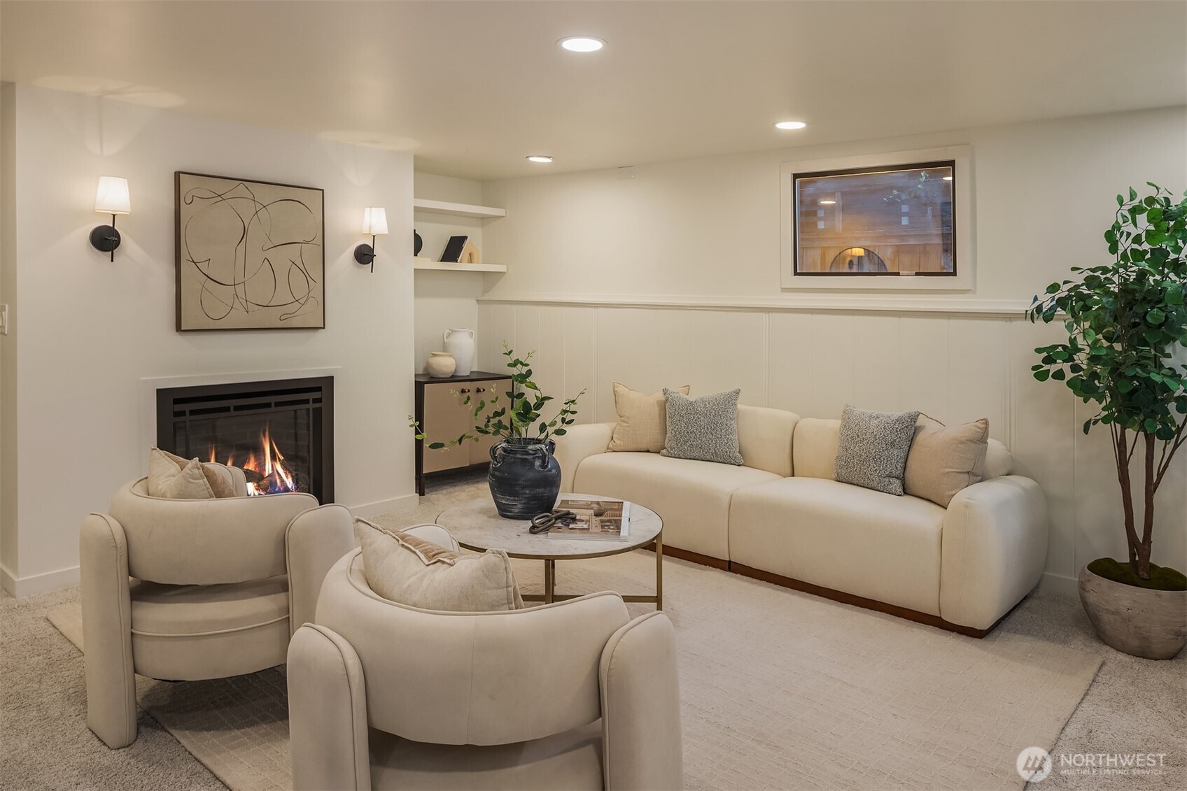 Staged living room with cream sofas and chairs around a coffee table beside a lit fireplace.
