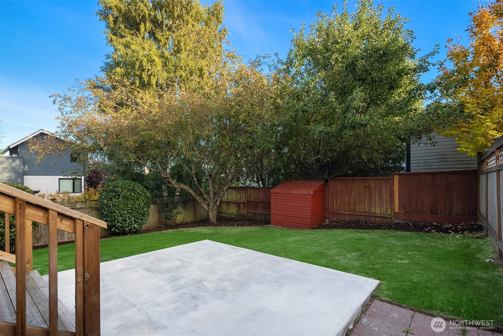 Fenced backyard with a concrete patio, green lawn, mature trees, and red storage shed.