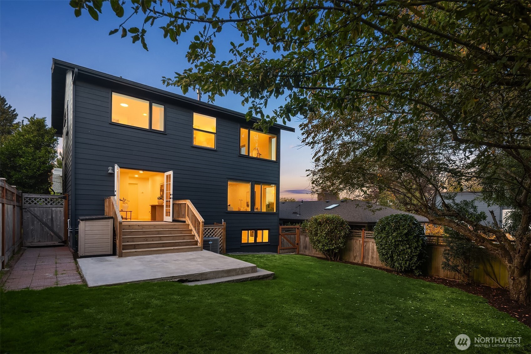 Modern dark-sided home with a warmly lit rear deck and lawn at dusk.