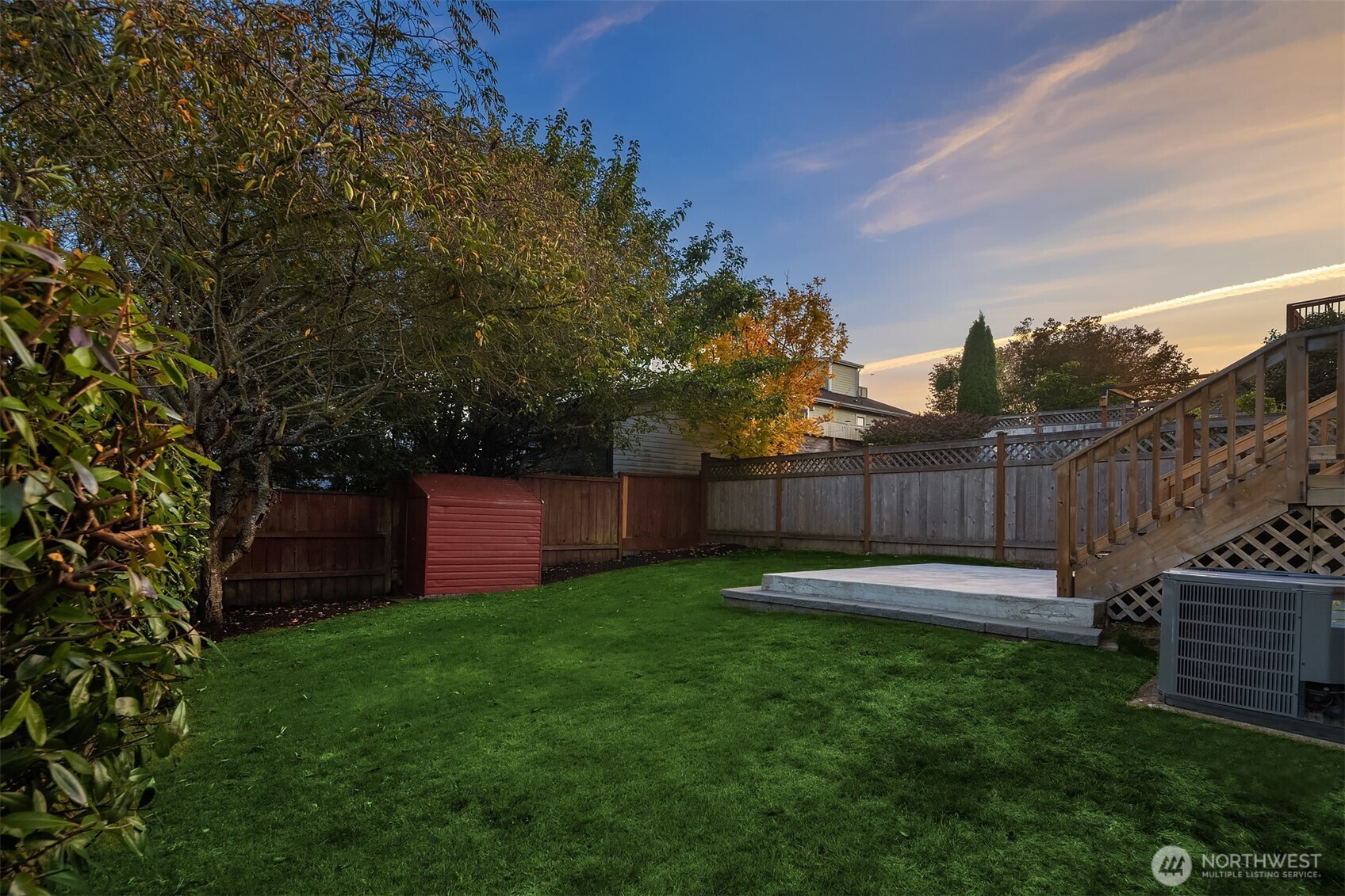 Fenced backyard with a green lawn, red shed, and raised wooden deck at sunset.
