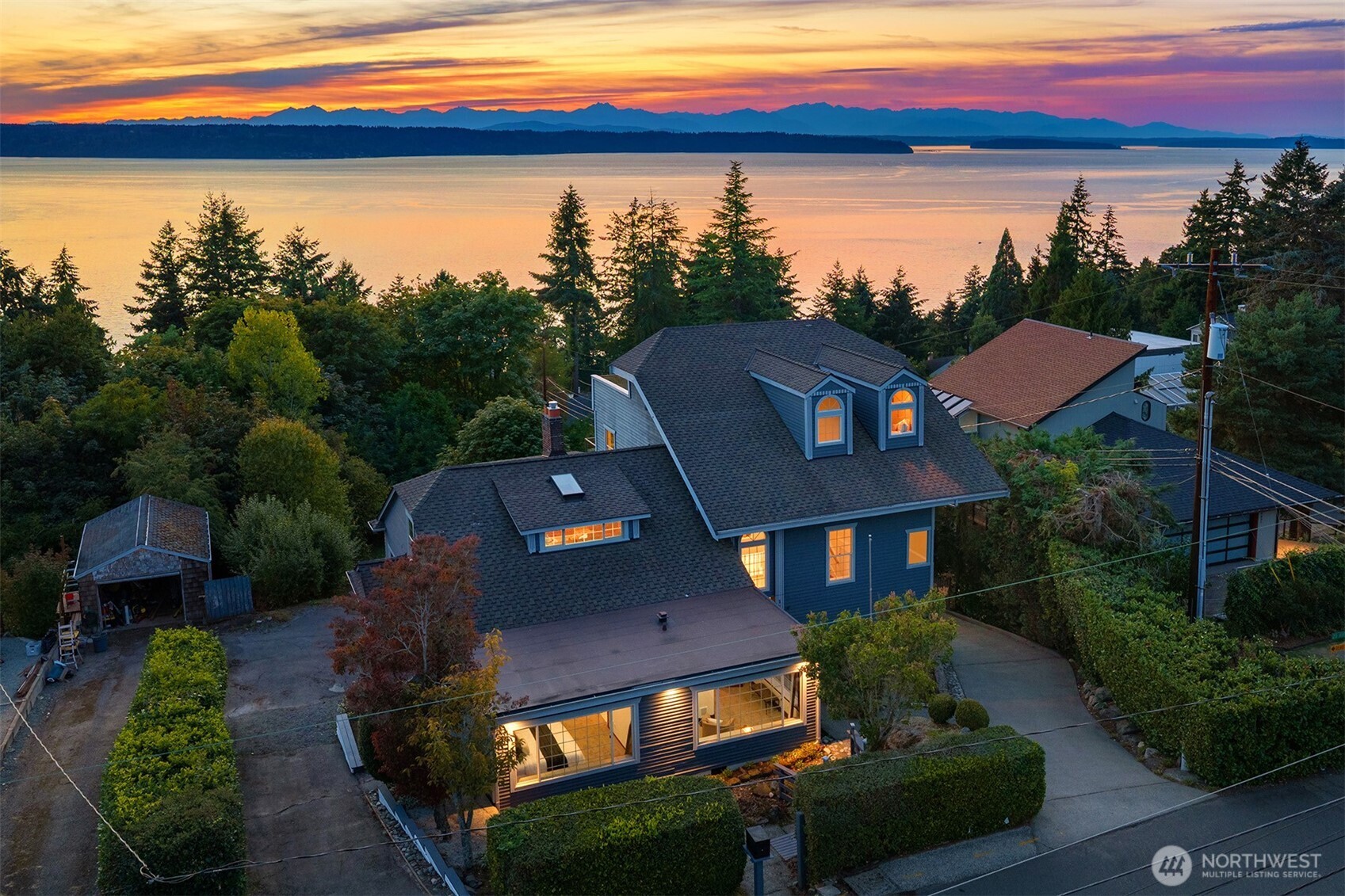 Aerial view of a blue home overlooking water and distant mountains at sunset.